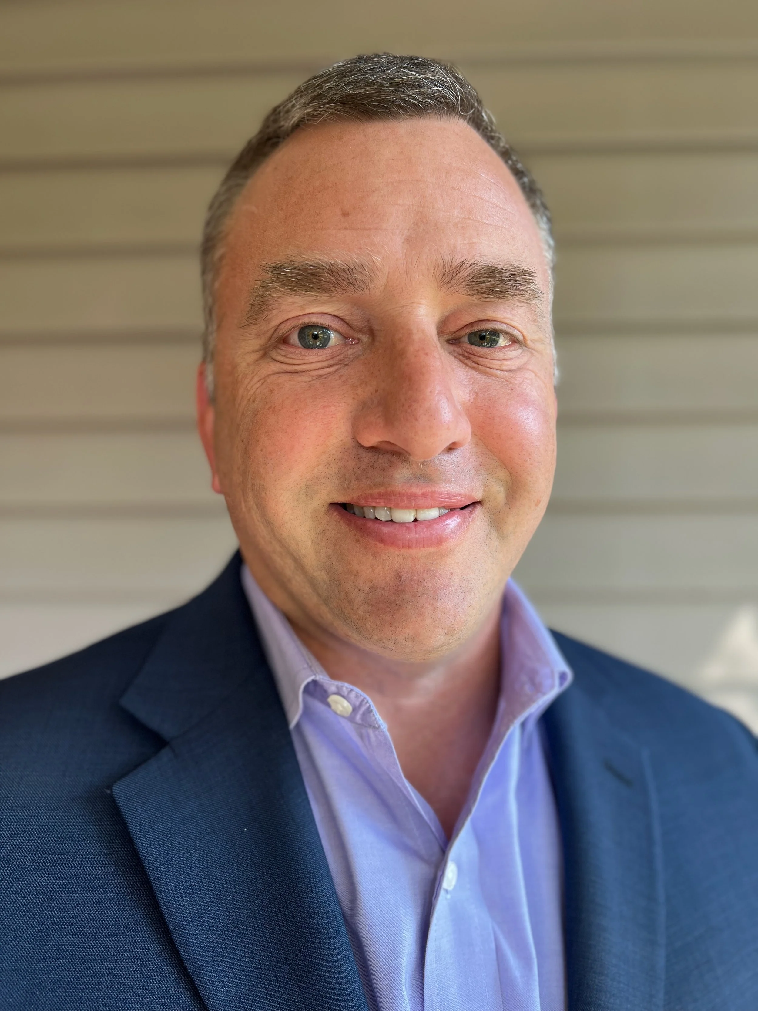 A man with short gray hair, bluish-green eyes, and light skin, dressed in a dark navy blazer and light purple dress shirt, standing against a beige, horizontally paneled background.