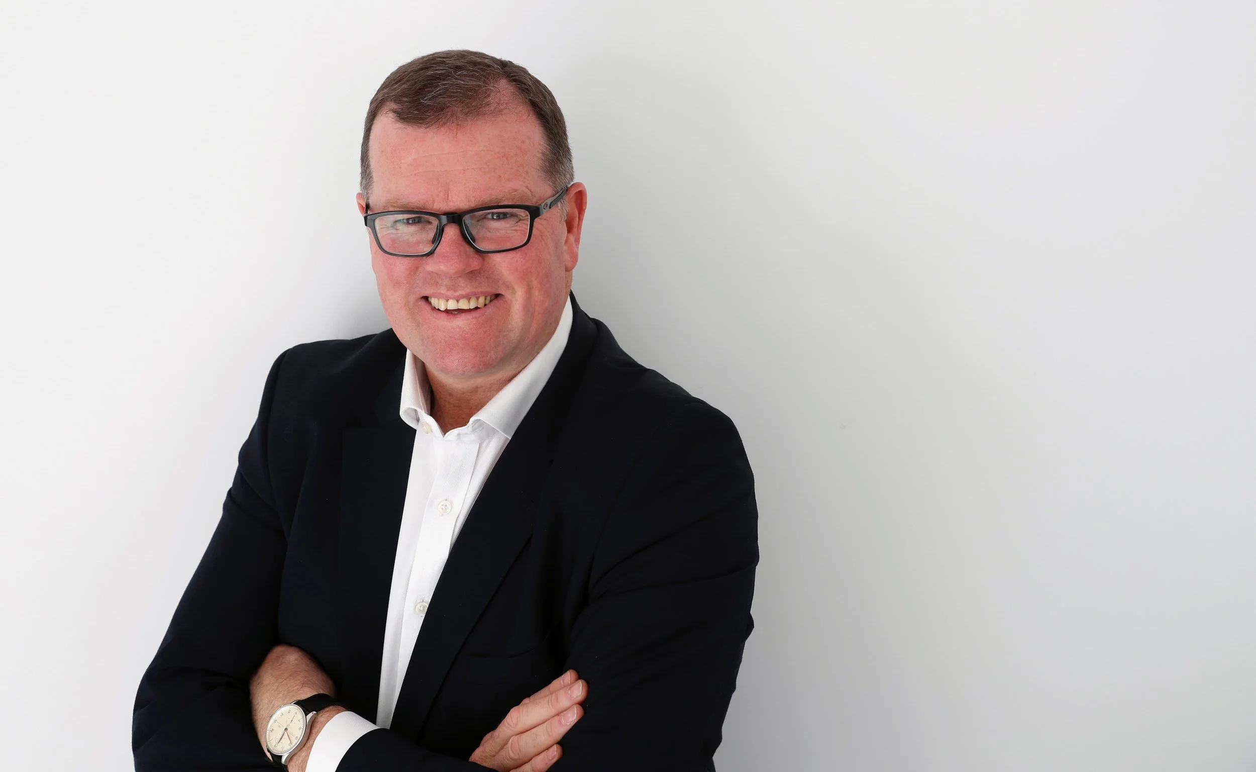A man with short gray hair, bluish-green eyes, and light skin, dressed in a dark navy blazer and light purple dress shirt, standing against a beige, horizontally paneled background.