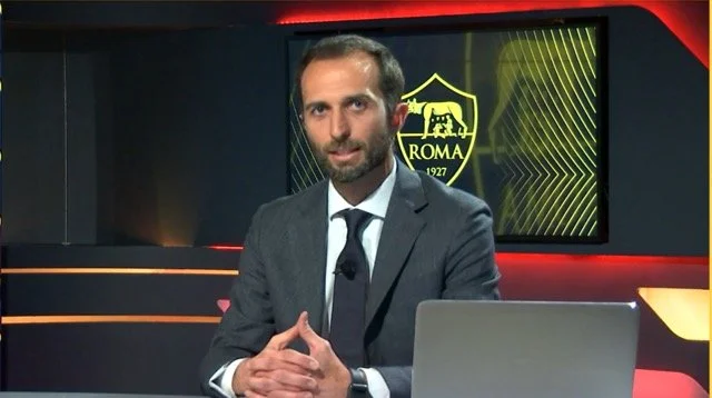 Man in a gray suit sitting at a desk with a laptop, in a studio with a screen displaying the AS Roma football club logo in the background.