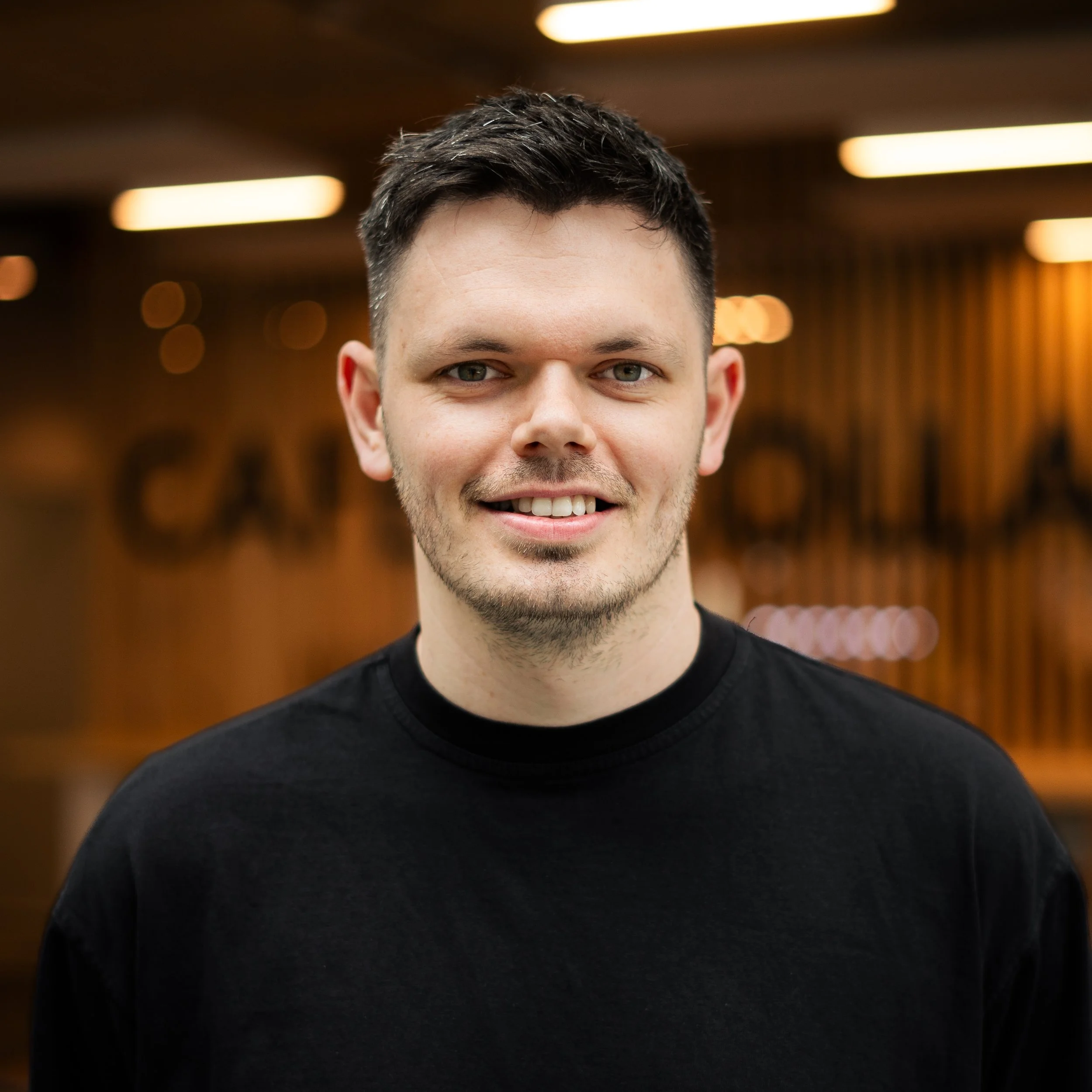 A young man with short dark hair and light stubble, smiling, wearing a black t-shirt, standing indoors with warm lighting and wooden wall decor.