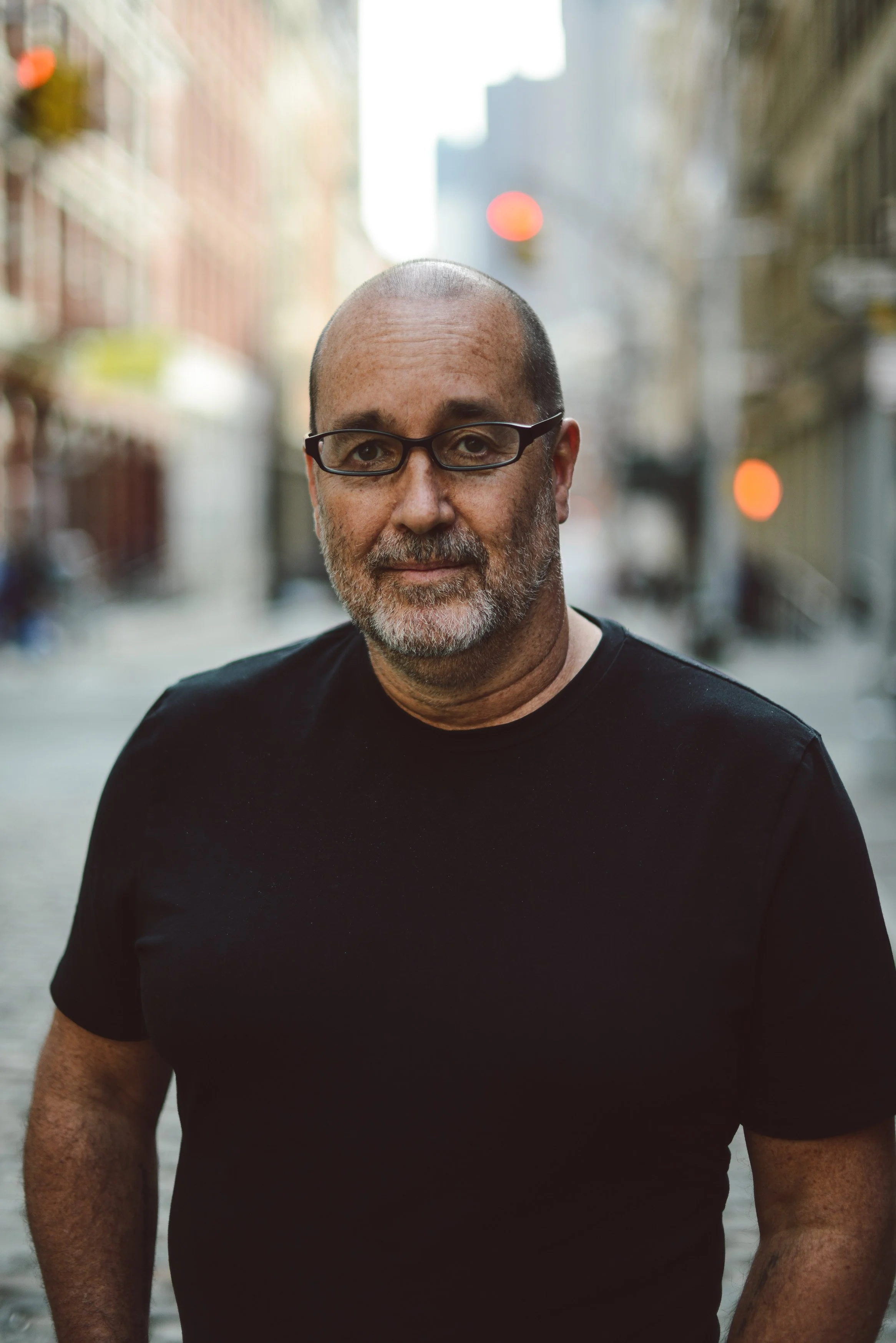 A middle-aged man with glasses, a beard, and a shaved head stands on a city street wearing a black t-shirt. The background features blurred buildings and streetlights.