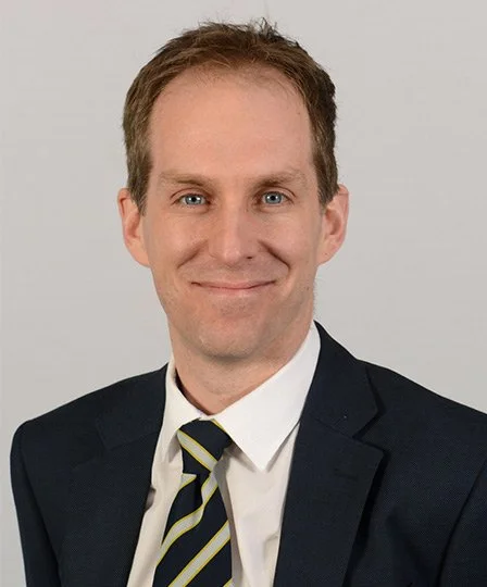 Professional headshot of a man in a dark suit, white shirt, and striped tie, smiling against a plain light background.