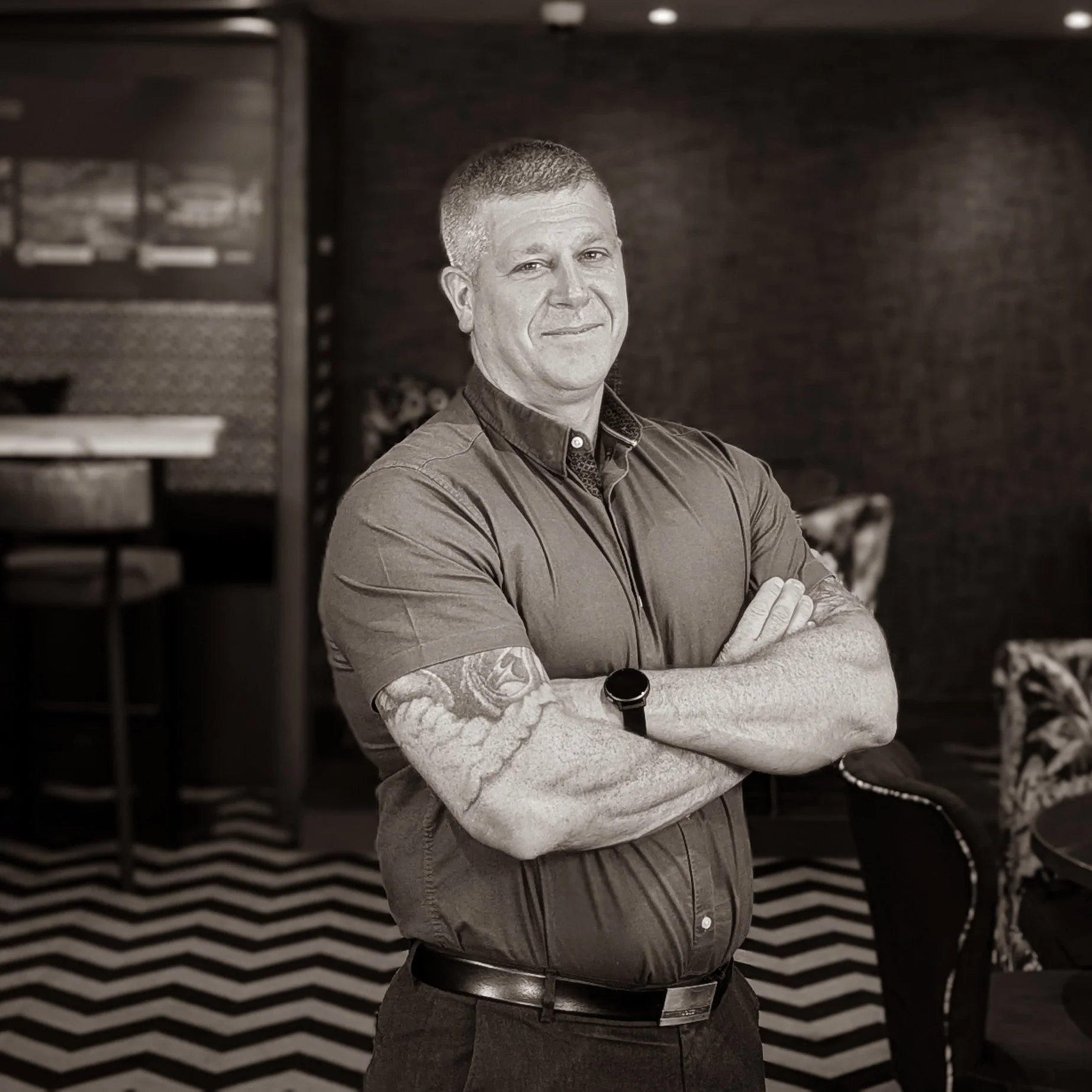 A man standing with arms crossed in a room with patterned carpet and dark brick walls.
