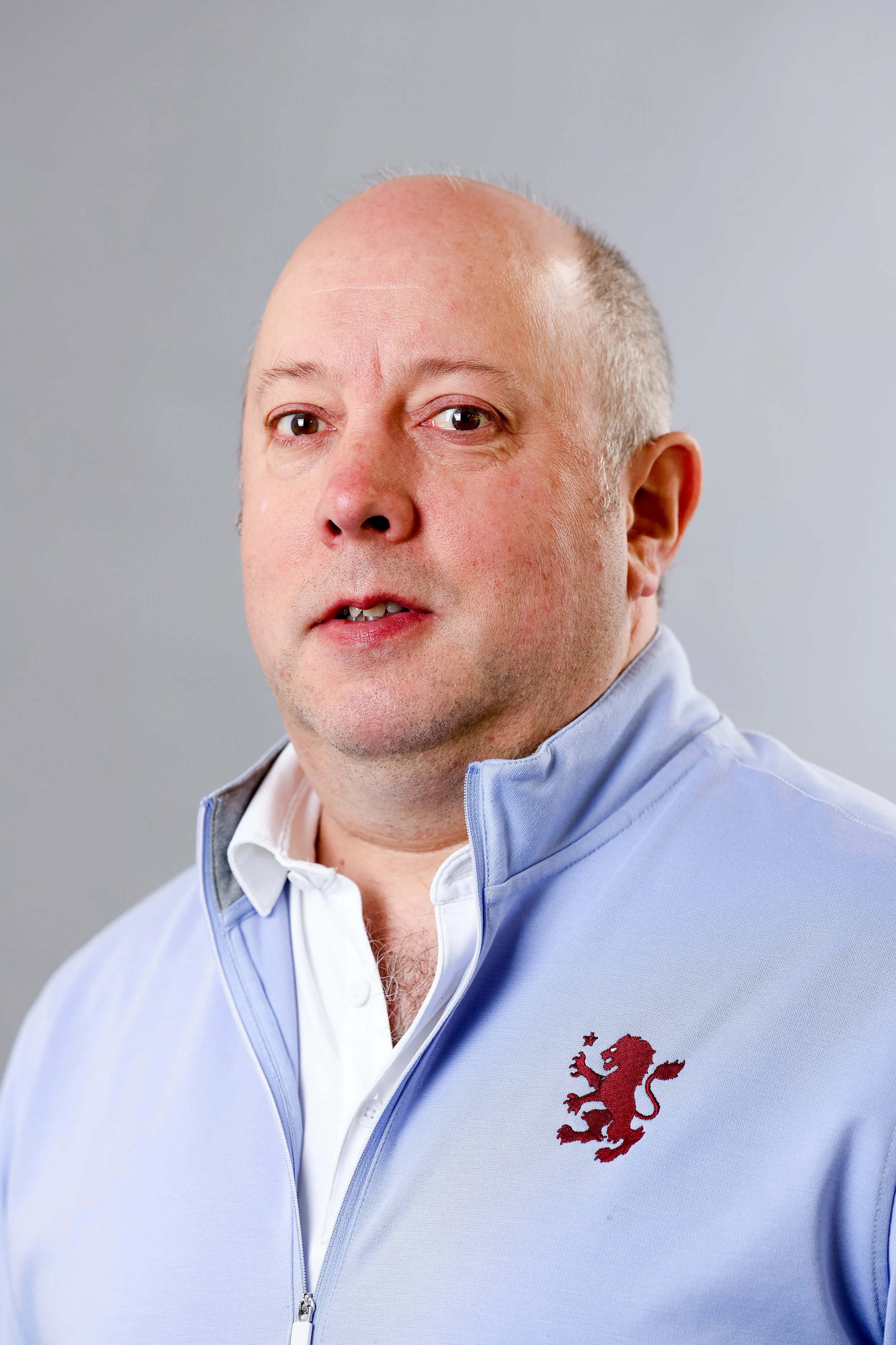 Portrait of a middle-aged man with a bald head, wearing a light blue jacket with a red lion emblem, against a gray background.
