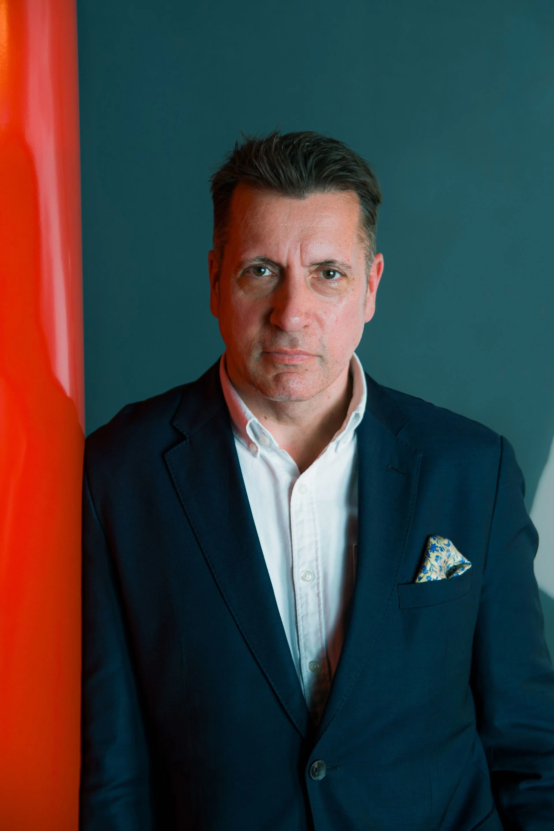 A man in a dark suit and white shirt standing indoors with a blue wall background, a red-orange object partially visible on the left, and a patterned pocket square.