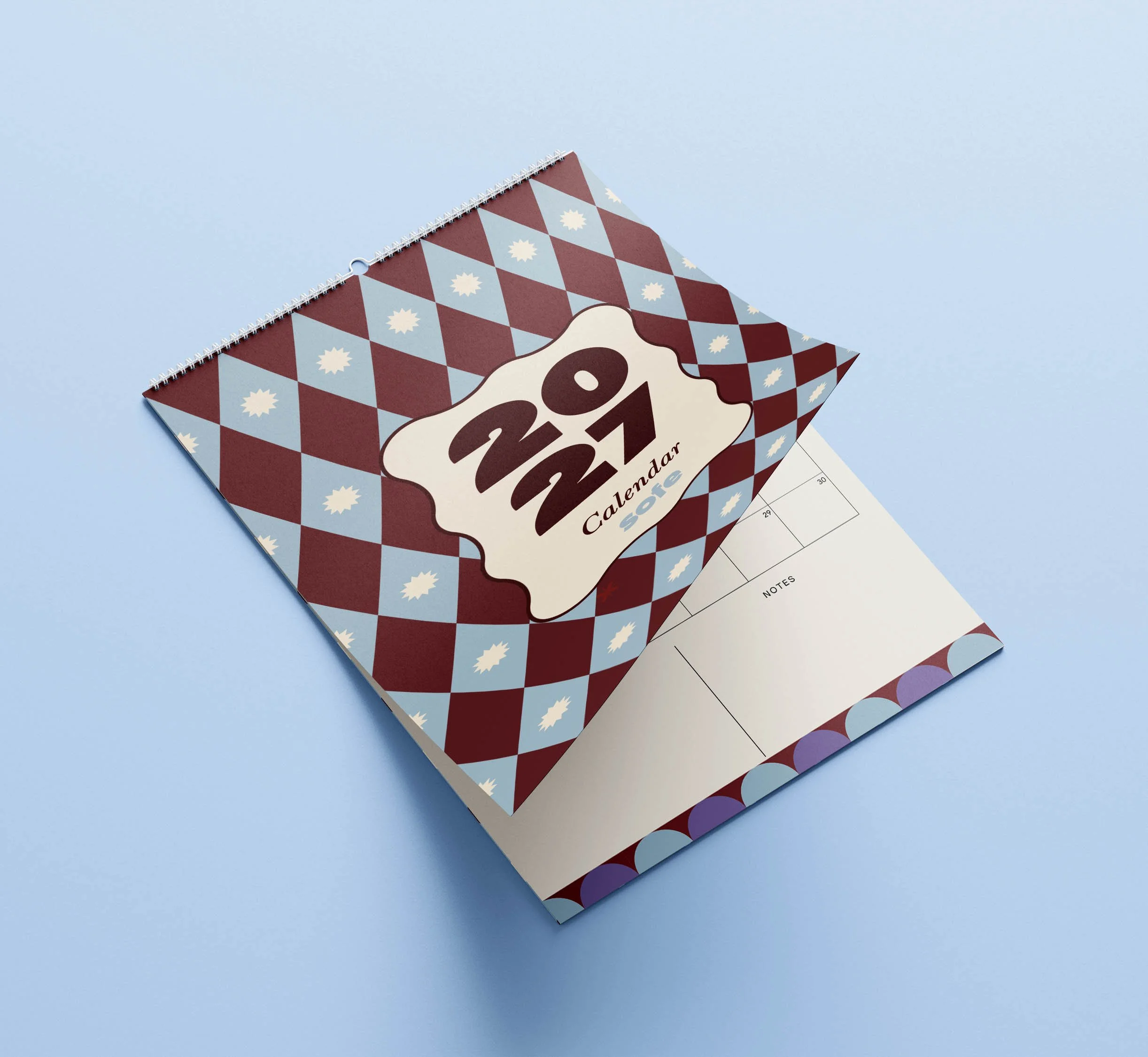 Sofe Store 2027 wall calendar lying at an angle on a pale blue background with the diamond pattern cover lifting to reveal the monthly planner page and Notes section beneath