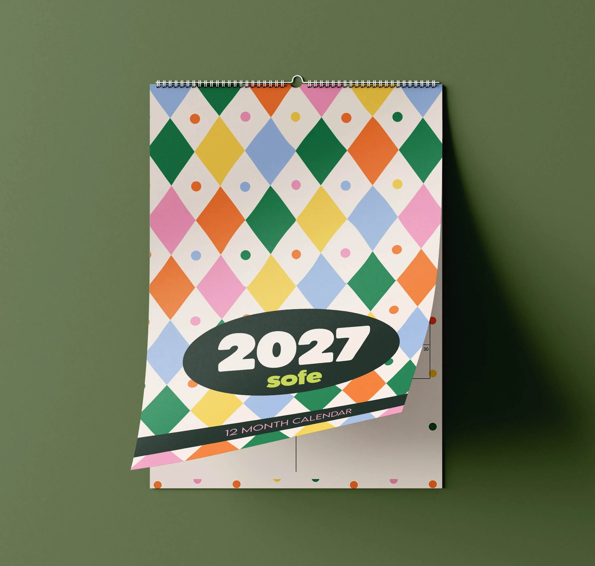 Sofe Store 2027 wall calendar with the harlequin pattern cover page curling up to reveal the monthly planner page beneath, on a green background