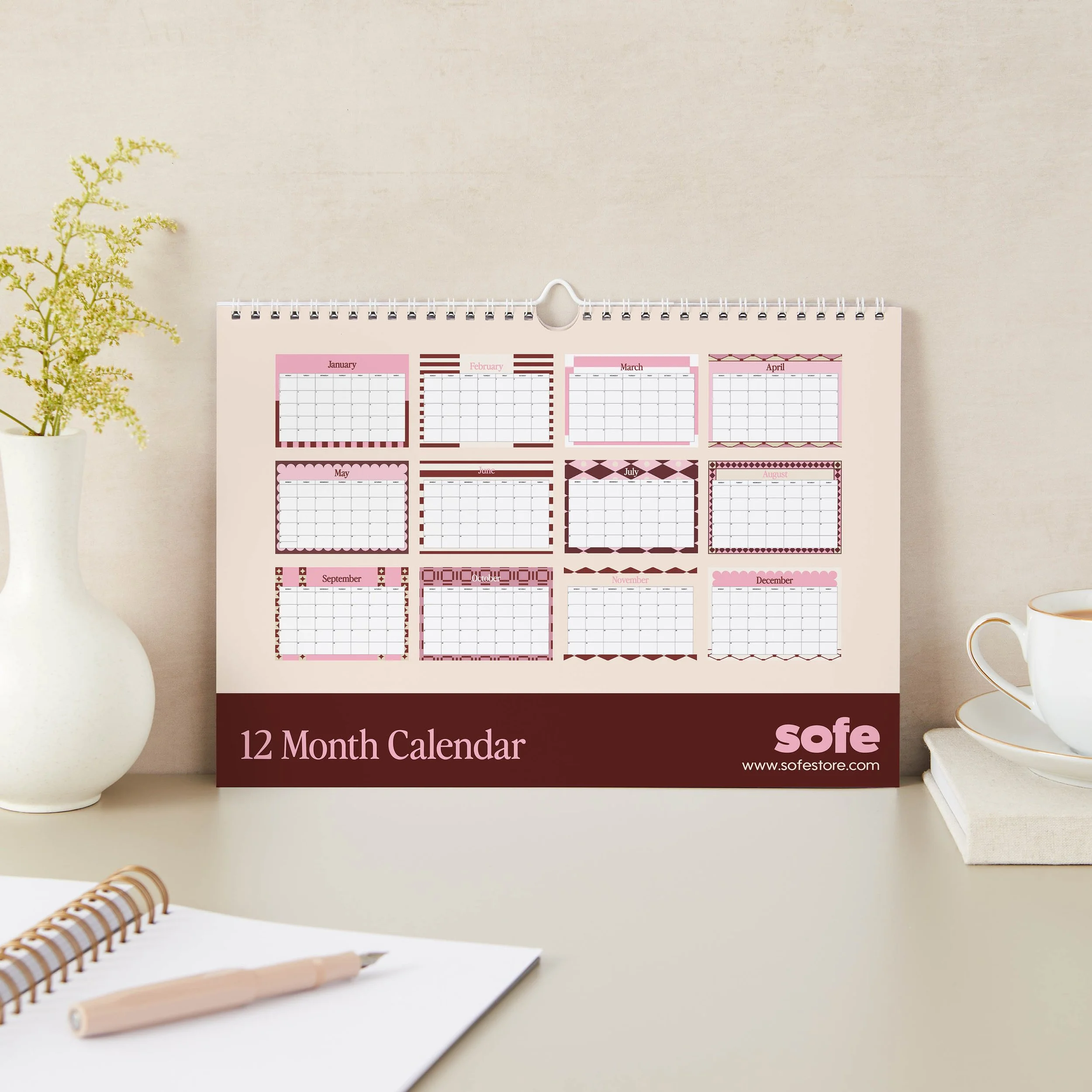 Back cover of a pink and burgundy 2027 landscape wall calendar showing a year-at-a-glance overview of all twelve monthly designs above a burgundy band reading 12 Month Calendar, on a neutral desk set