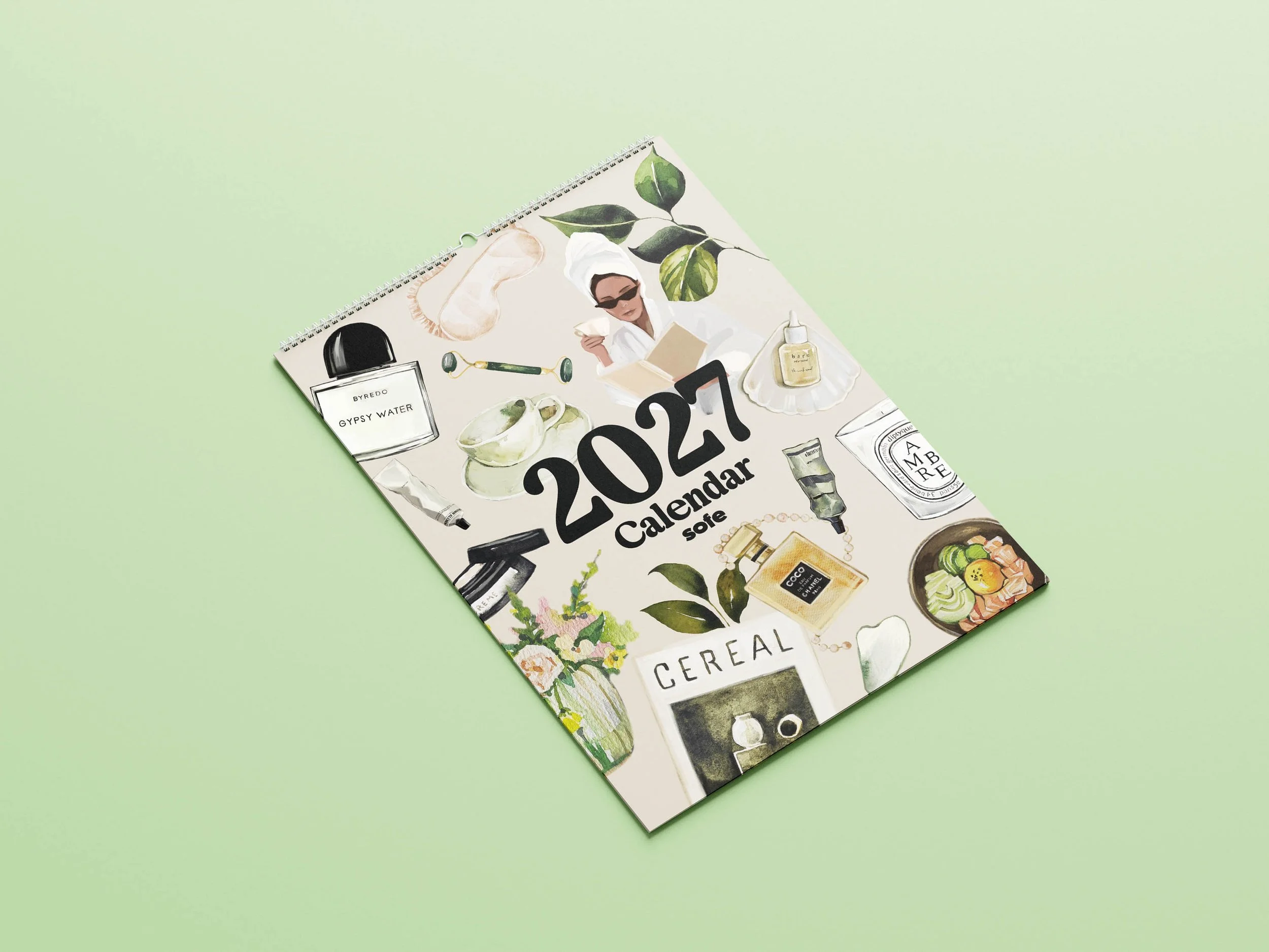 Sofe Store 2027 wall calendar lying at an angle on a green background, showing the watercolour wellness cover with skincare, a scented candle, fresh flowers and the “2027 Calendar” title