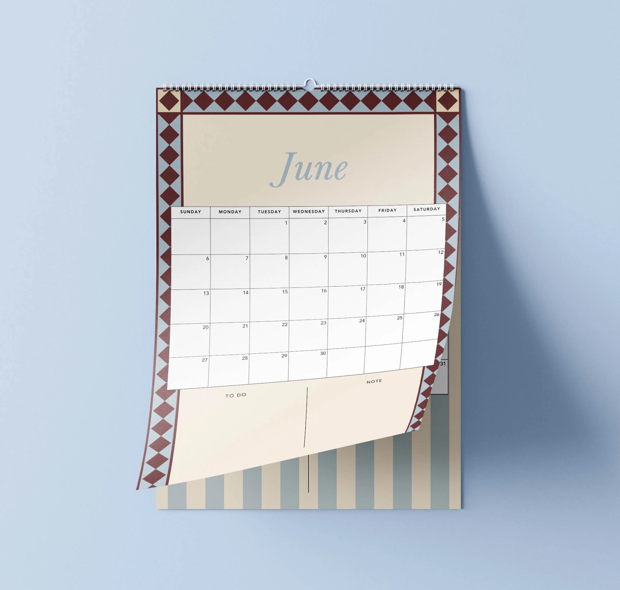 June 2027 page of the Sofe Store wall calendar curling up, with a burgundy and blue diamond border, a dusty blue “June” heading, a Sunday-to-Saturday date grid and To Do and Notes sections