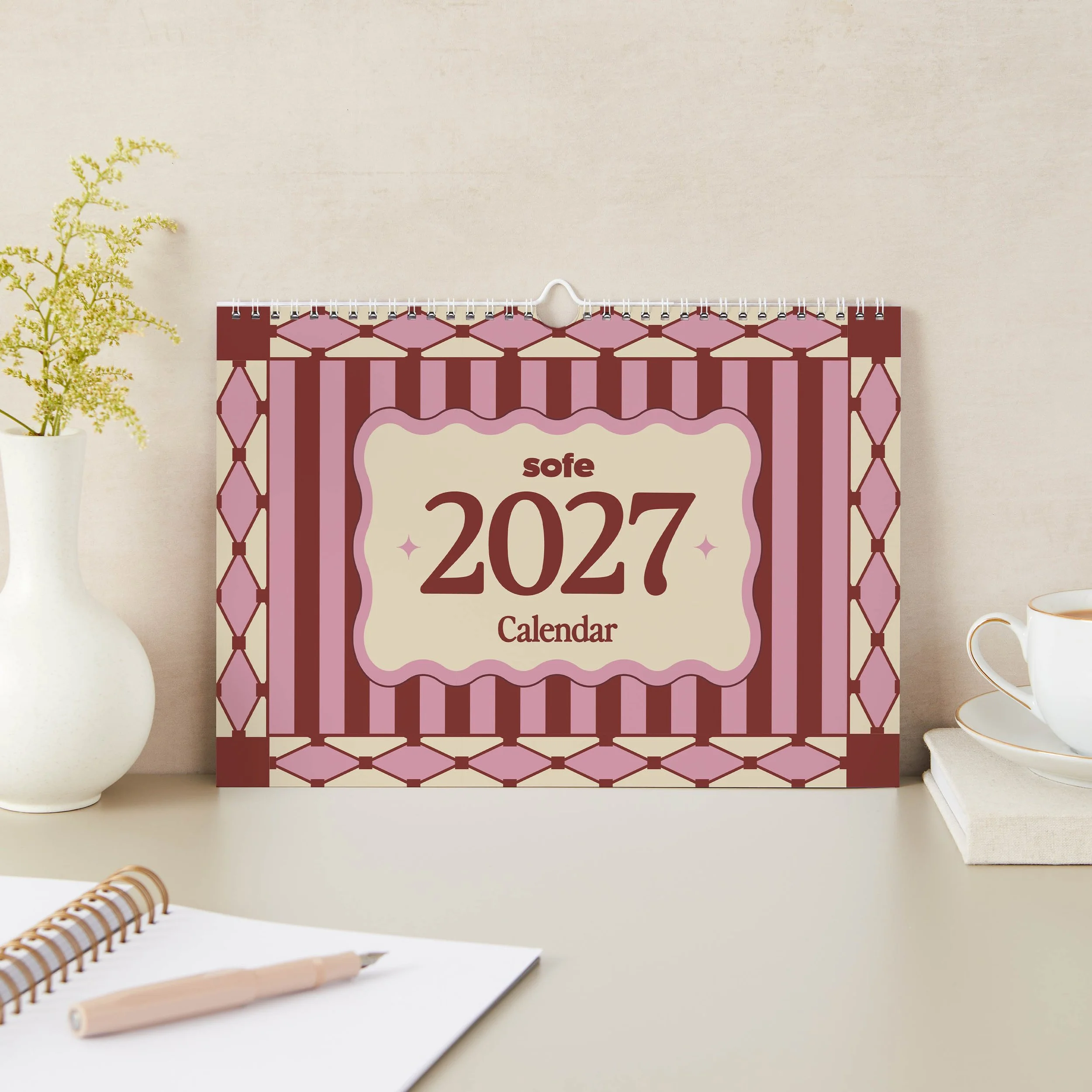 2027 landscape wall calendar cover with pink and burgundy stripes, harlequin diamond borders and a wavy cream label reading 2027 Calendar, standing on a desk beside a white vase, teacup and notebook