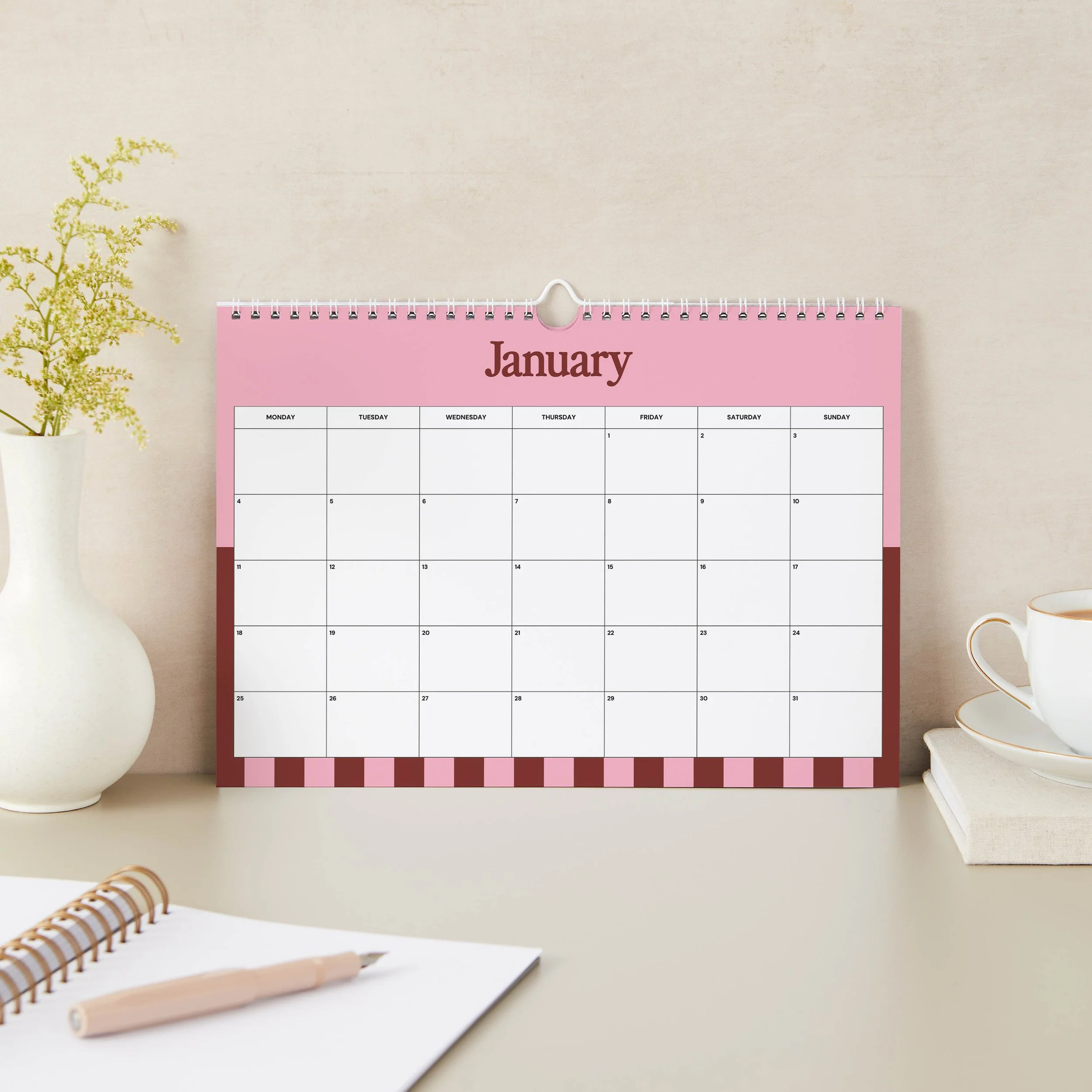January 2027 wall calendar page with a pink header, burgundy side panels, a candy-striped bottom border and a large Monday-start date grid, styled with a white vase and notebook