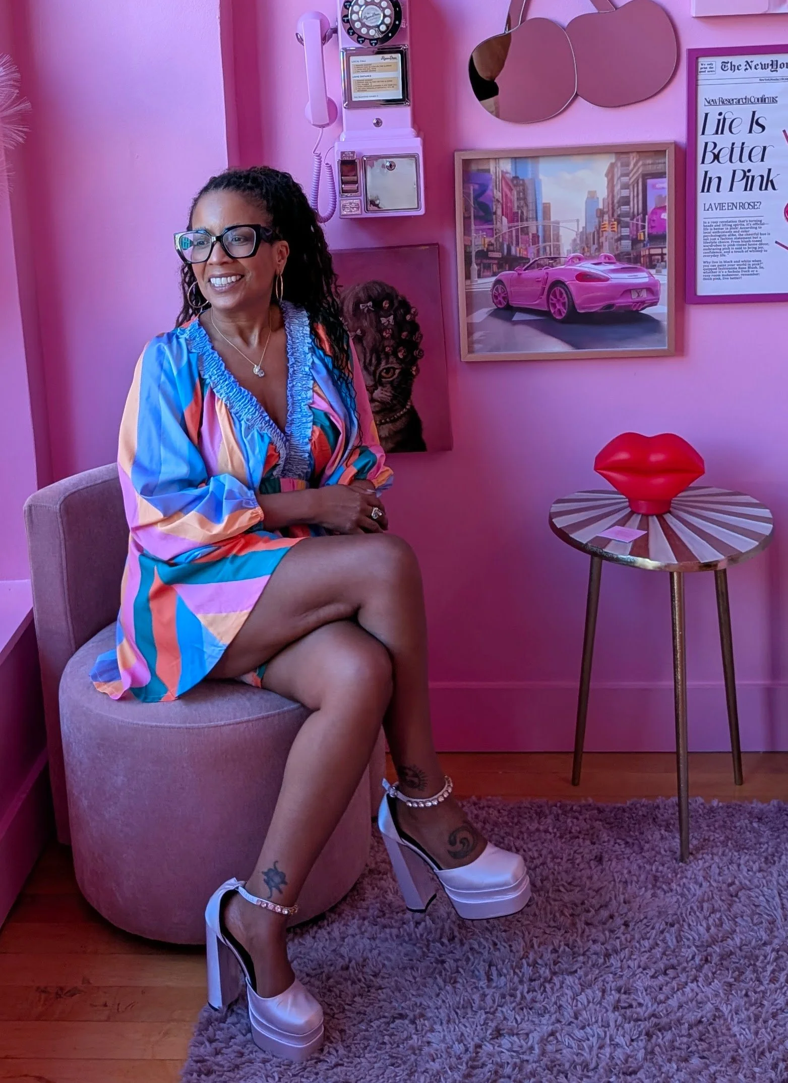 A woman sitting on a pink cushioned stool in a room with pink walls, wearing a colorful striped dress and high platform shoes, smiling and looking to her left. The room has wall art, including mirrors, a framed cityscape picture, and a poster with pink-themed content. There is also a small table with a large red lips sculpture on it.