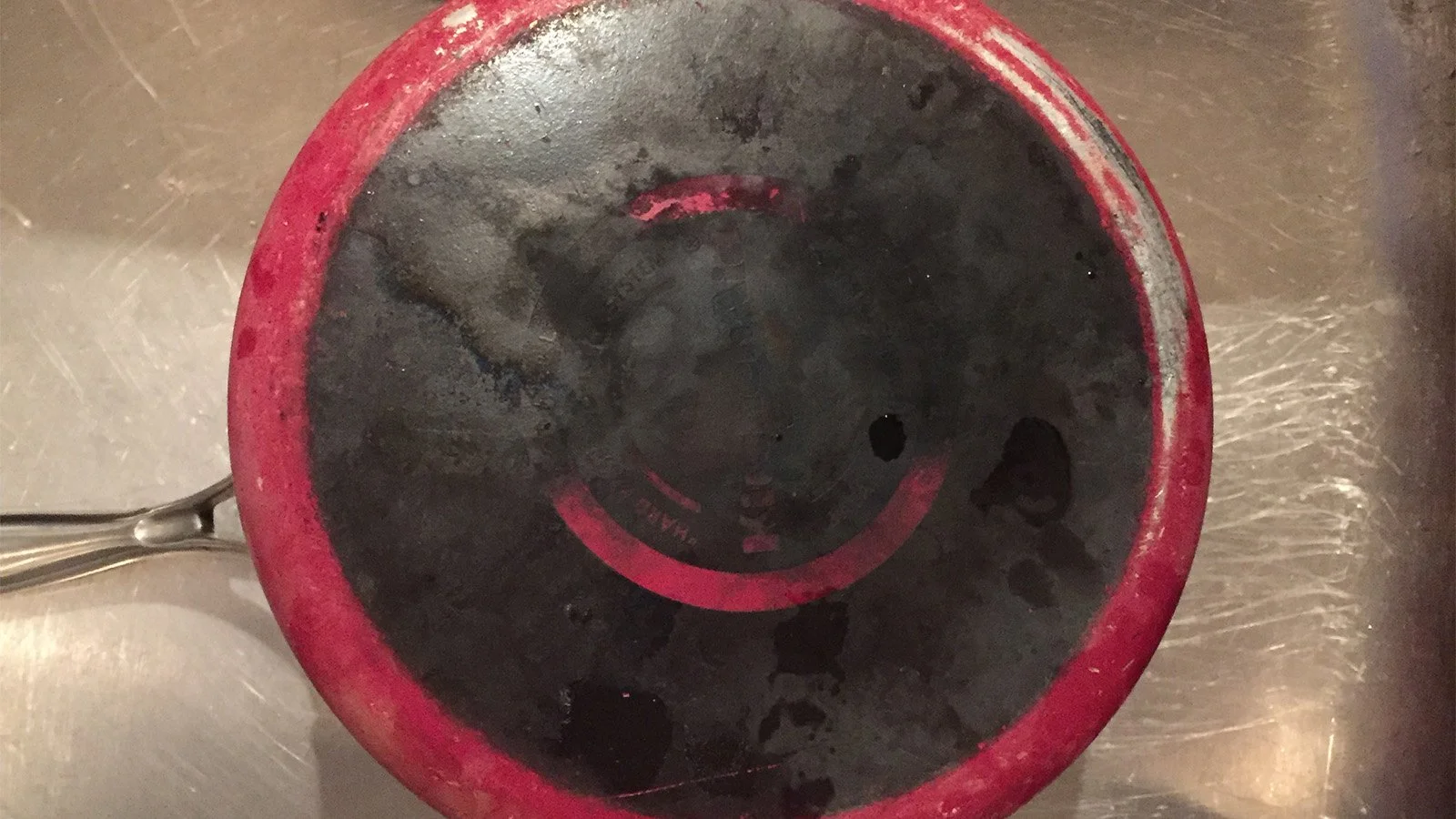 A red pan on a stove with black charred and burnt residue inside, indicating it has been used to cook something that burned or overcooked.