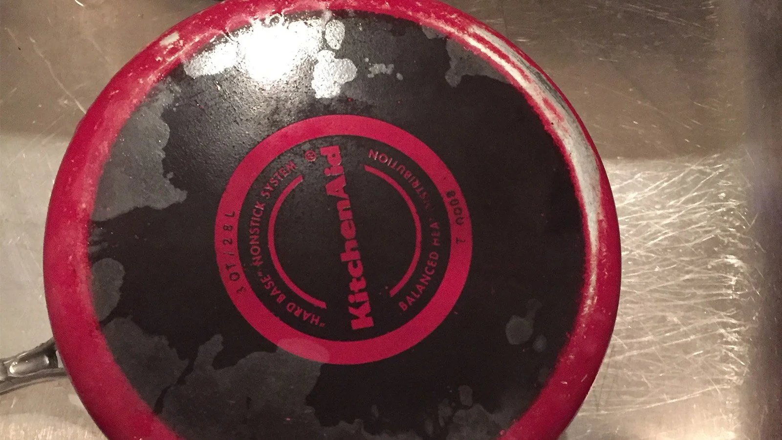 A close-up view of the bottom of a red and black cast iron pan showing the brand name 'KitchenAid' and text indicating it is a 'Handcast in Monstic' with a capacity of 1.2 liters.