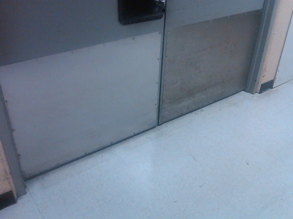 Close-up of metal classroom or office door with a black door handle, situated on a light gray concrete or linoleum floor.