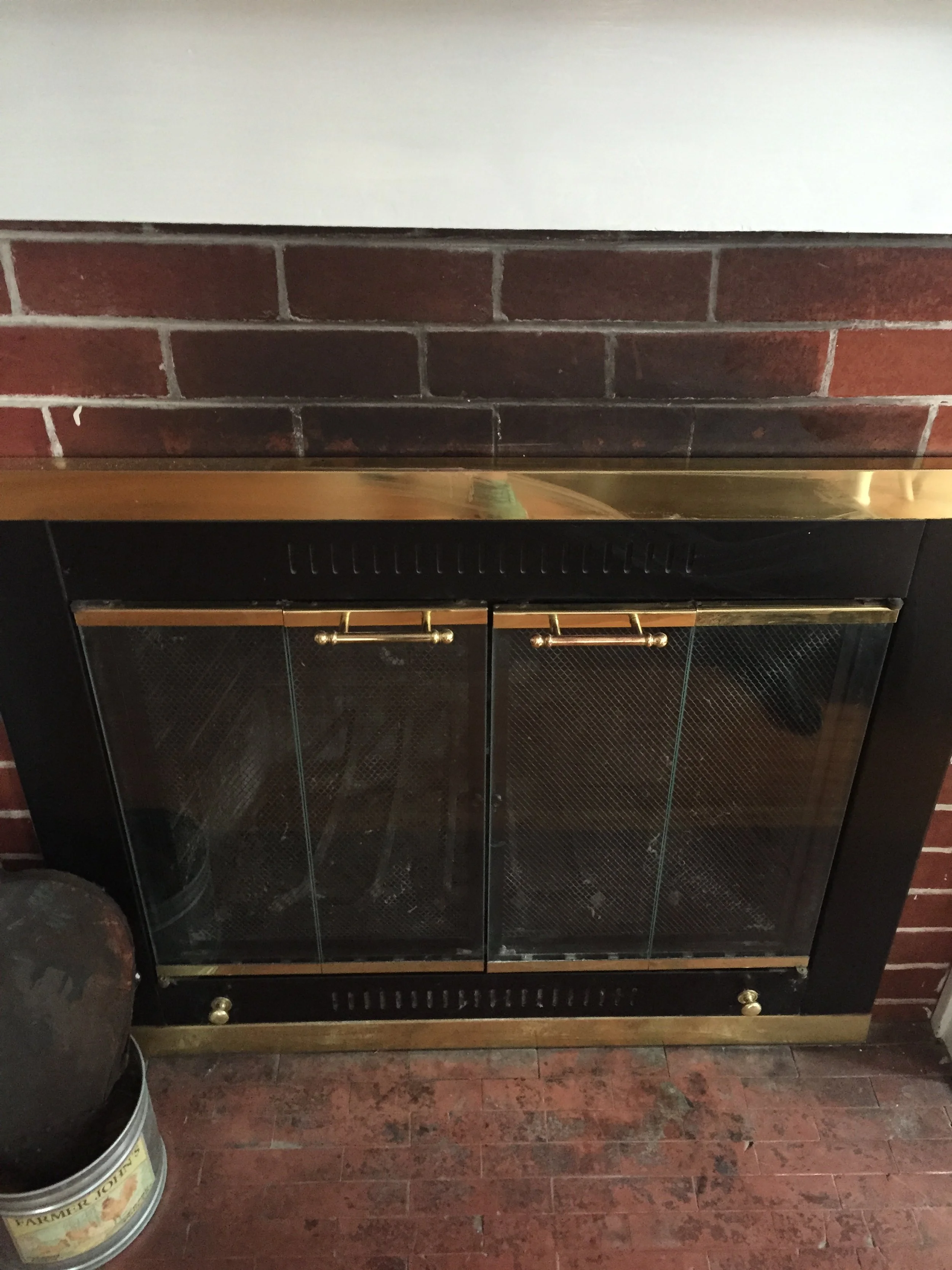 A black and gold cabinet with glass doors and a wooden top, set on a brick floor, with a metal container of paint or similar product nearby.