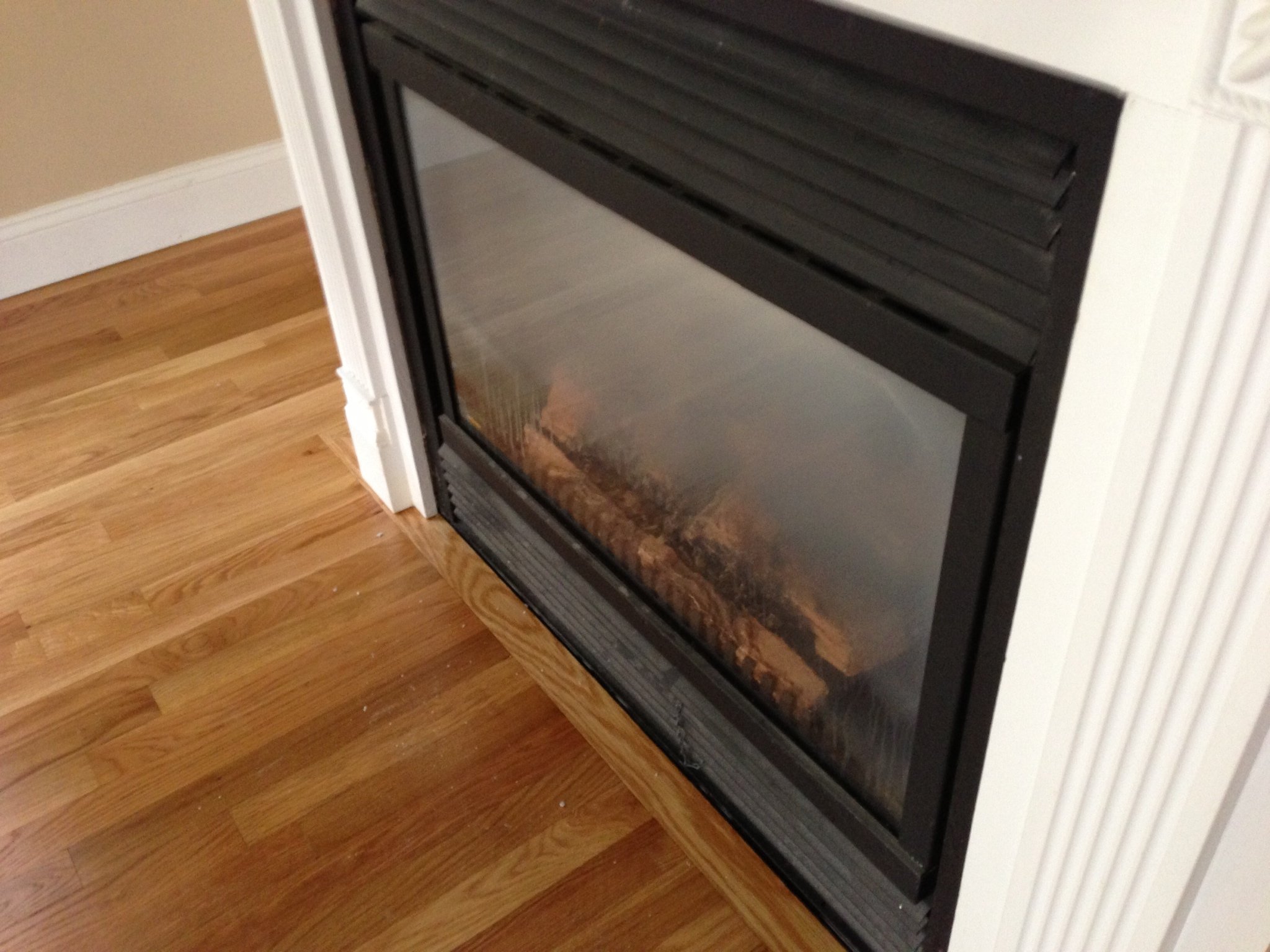 A fireplace with a black metal frame and glass front, set into a white wall, with a hardwood floor nearby.