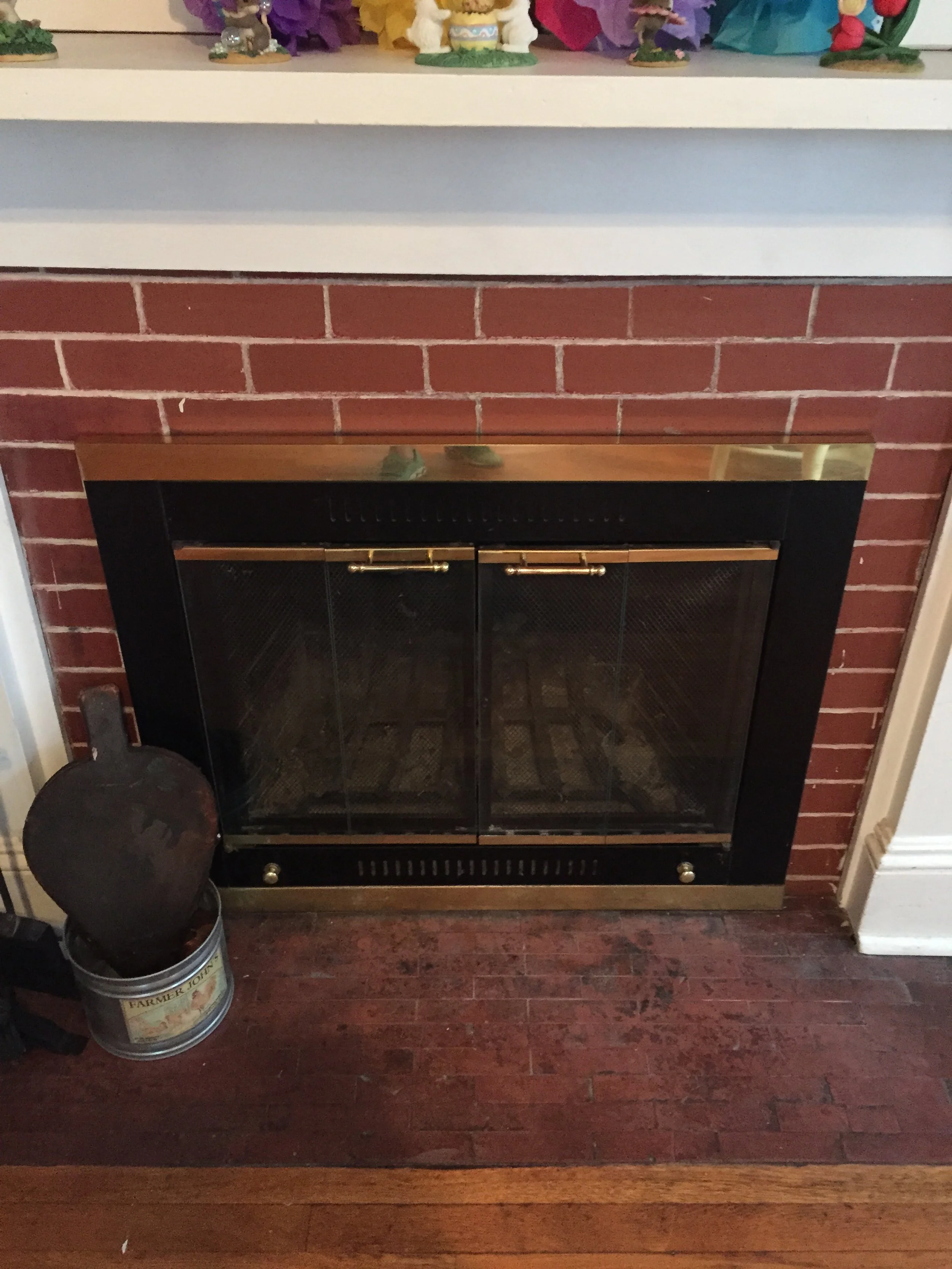 A fireplace with a brick surround and black finish, with gold accents on the hearth. A metal bucket with a fire poker and a round metal item are placed to the left of the fireplace. The mantel has colorful decorative items on top, including small fig