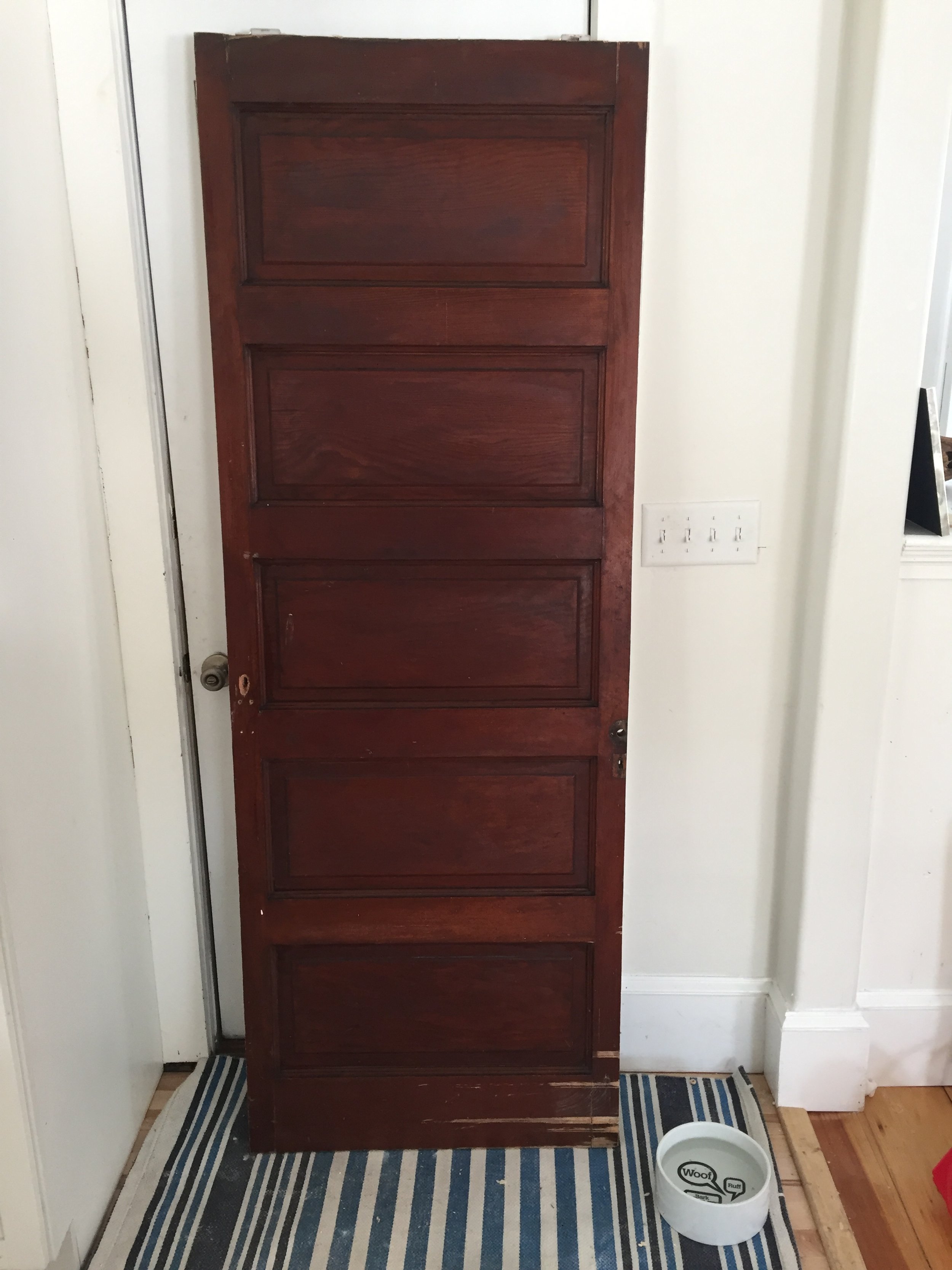 A tall wooden cabinet with three drawers and a small black doorknob on the right is placed against a white wall near a door and a white electrical outlet. A striped rug lies on the wooden floor in front of the cabinet. To the right of the cabinet is 
