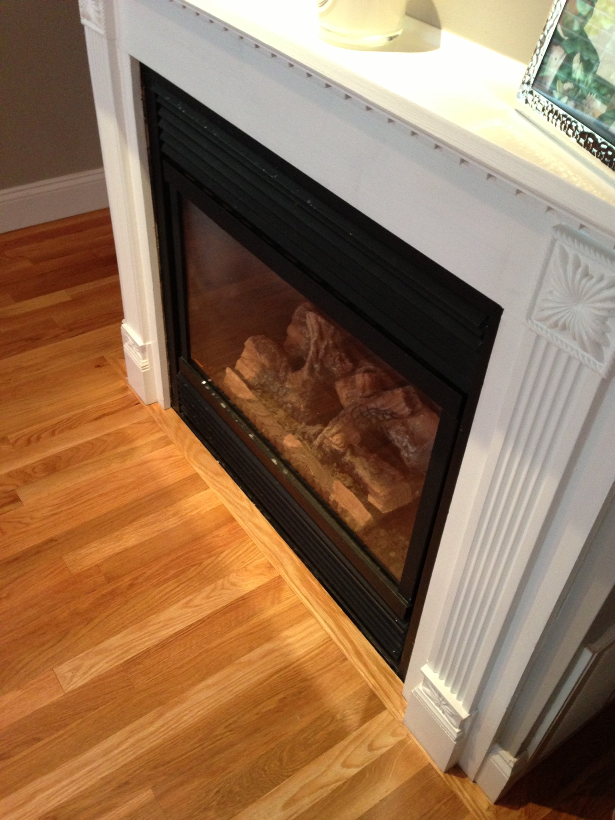 Wooden floor with a fireplace that has a glass front and a black metal frame, decorated with stone details on the mantel, next to a white radiator.