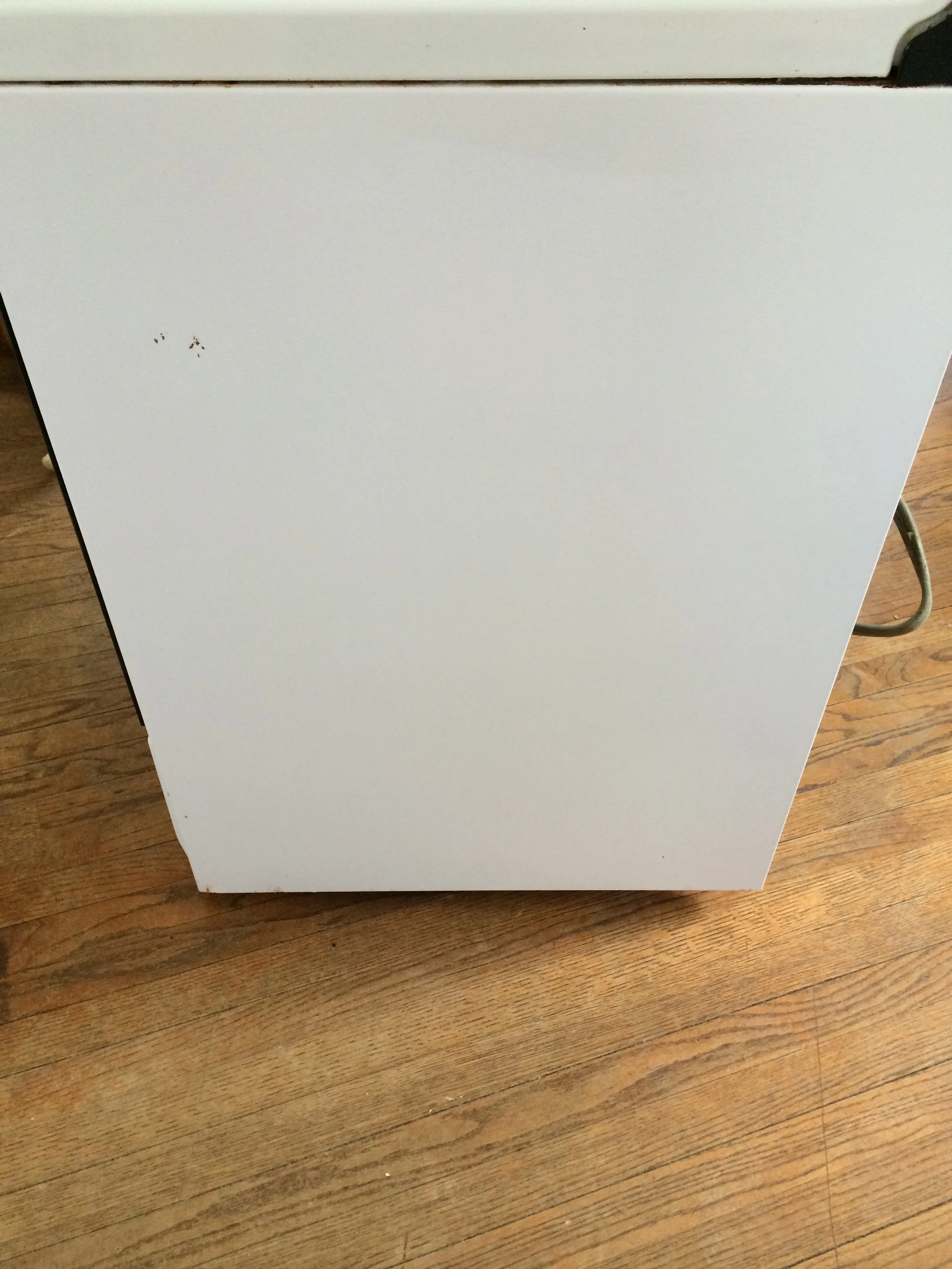 Close-up of a white appliance, possibly a dishwasher or refrigerator, on a wooden floor with a power cord visible on the right side.