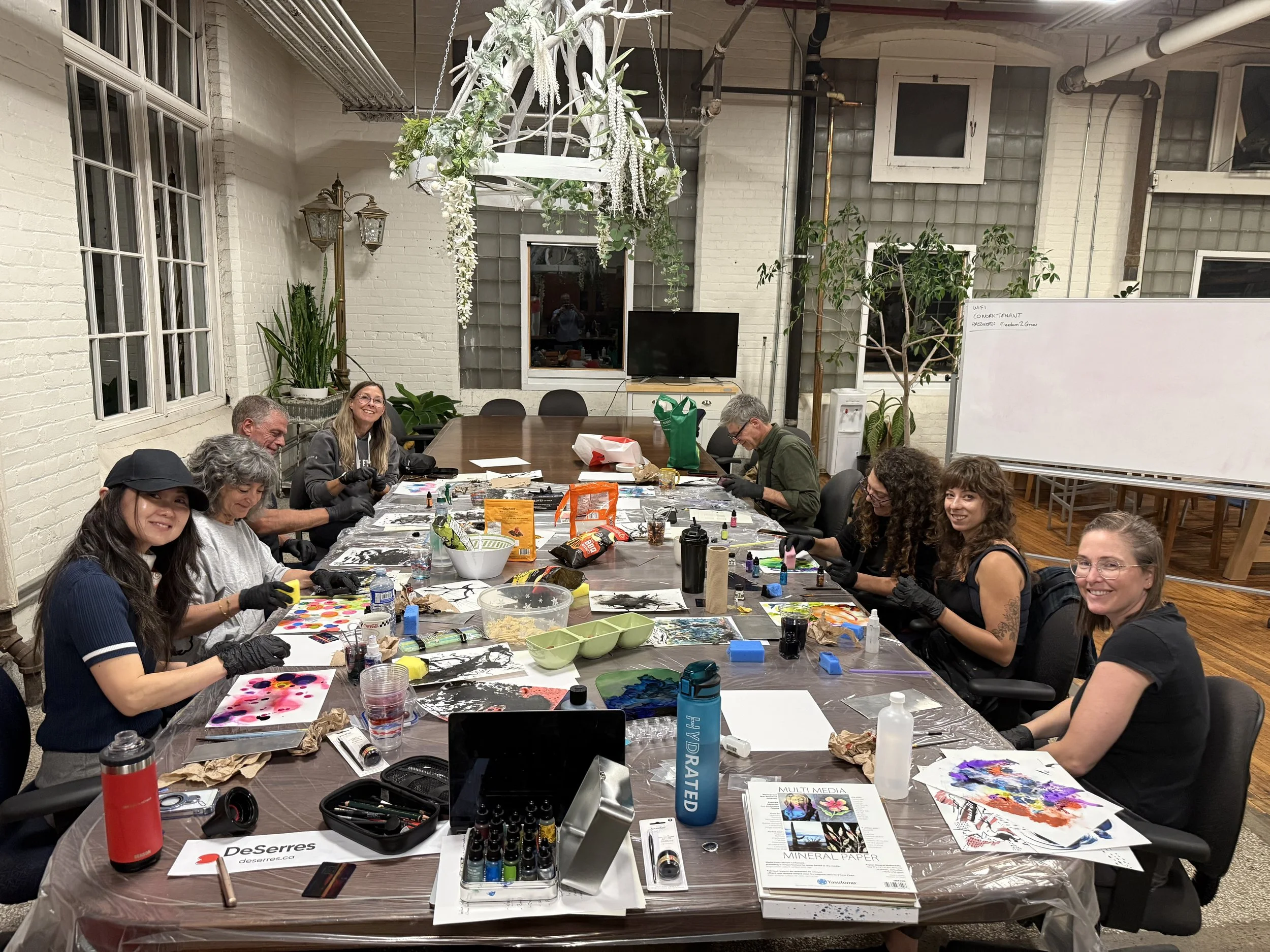 People sitting around a large table engaged in an art or crafting activity, with various supplies such as paints, brushes, and paper spread across the table in a well-lit room with white brick walls and large windows.