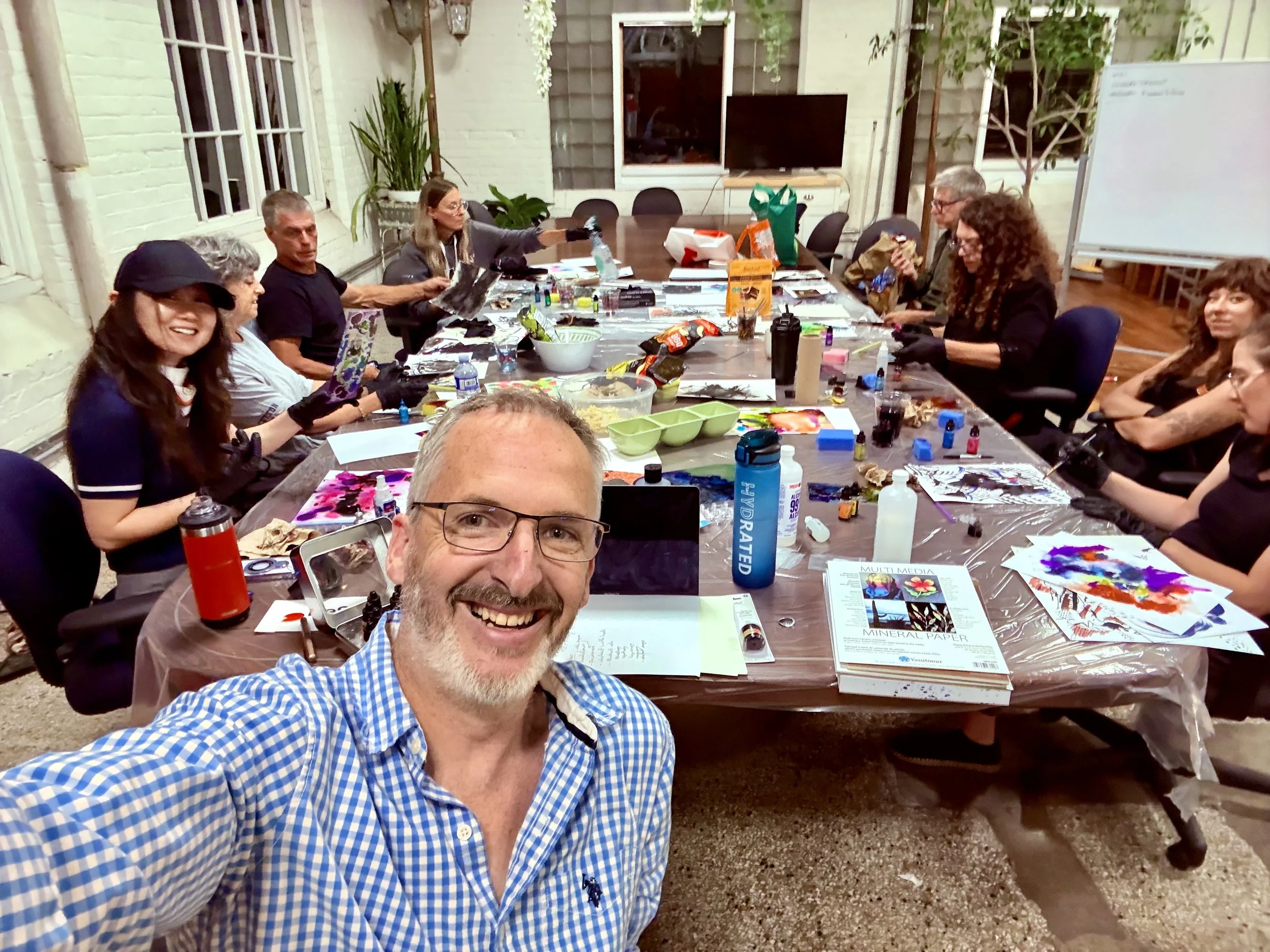 A group of people gathered around a large table, engaging in a painting workshop, with art supplies scattered on the table, and a man taking a selfie in the foreground.
