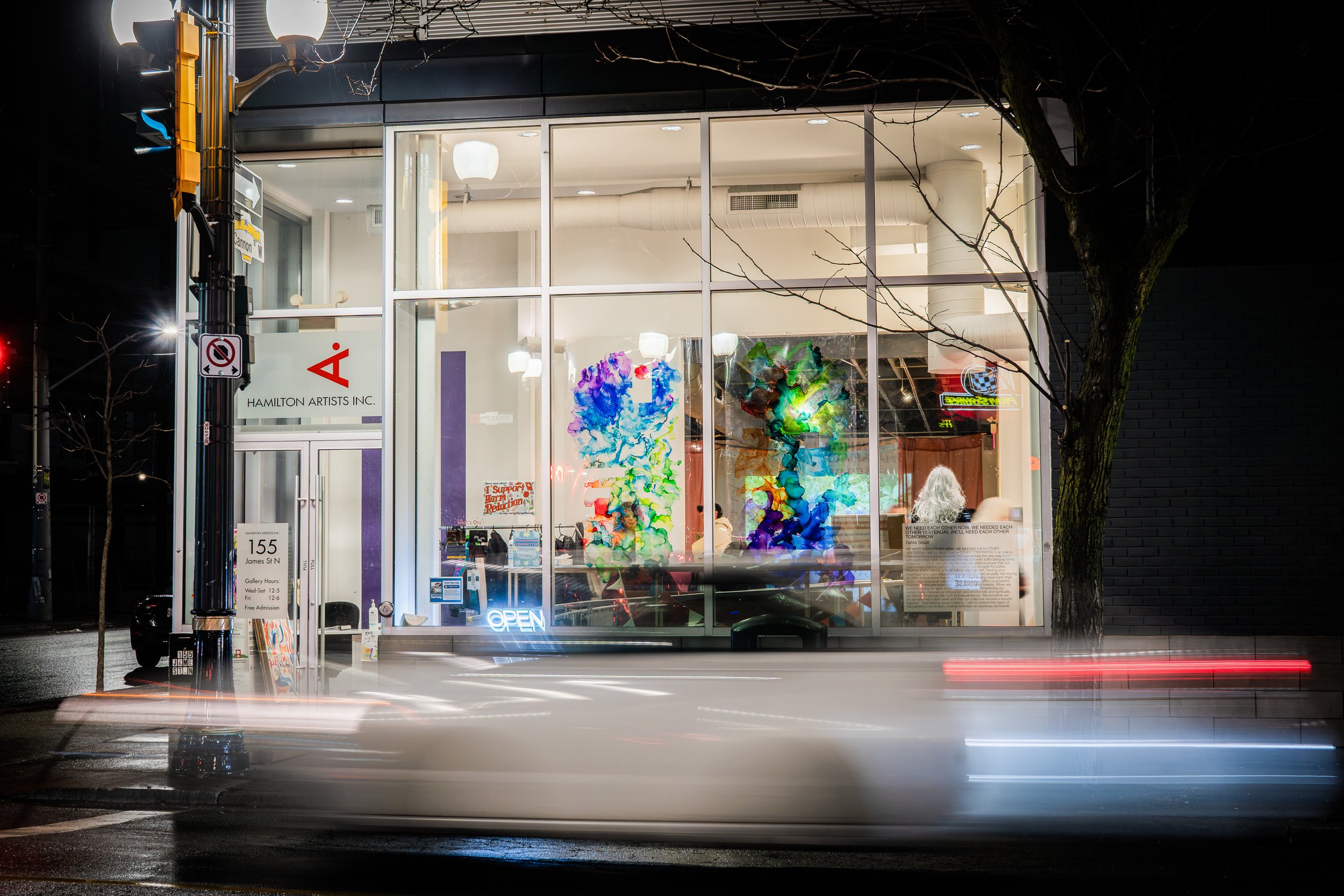 Nighttime view of Hamilton Artists Inc. art gallery with large glass windows displaying colorful abstract paintings inside. The gallery is illuminated, and a neon 'Open' sign is visible. A street scene with light streaks from passing vehicles is in the foreground, and a tree and streetlamp are near the building.
