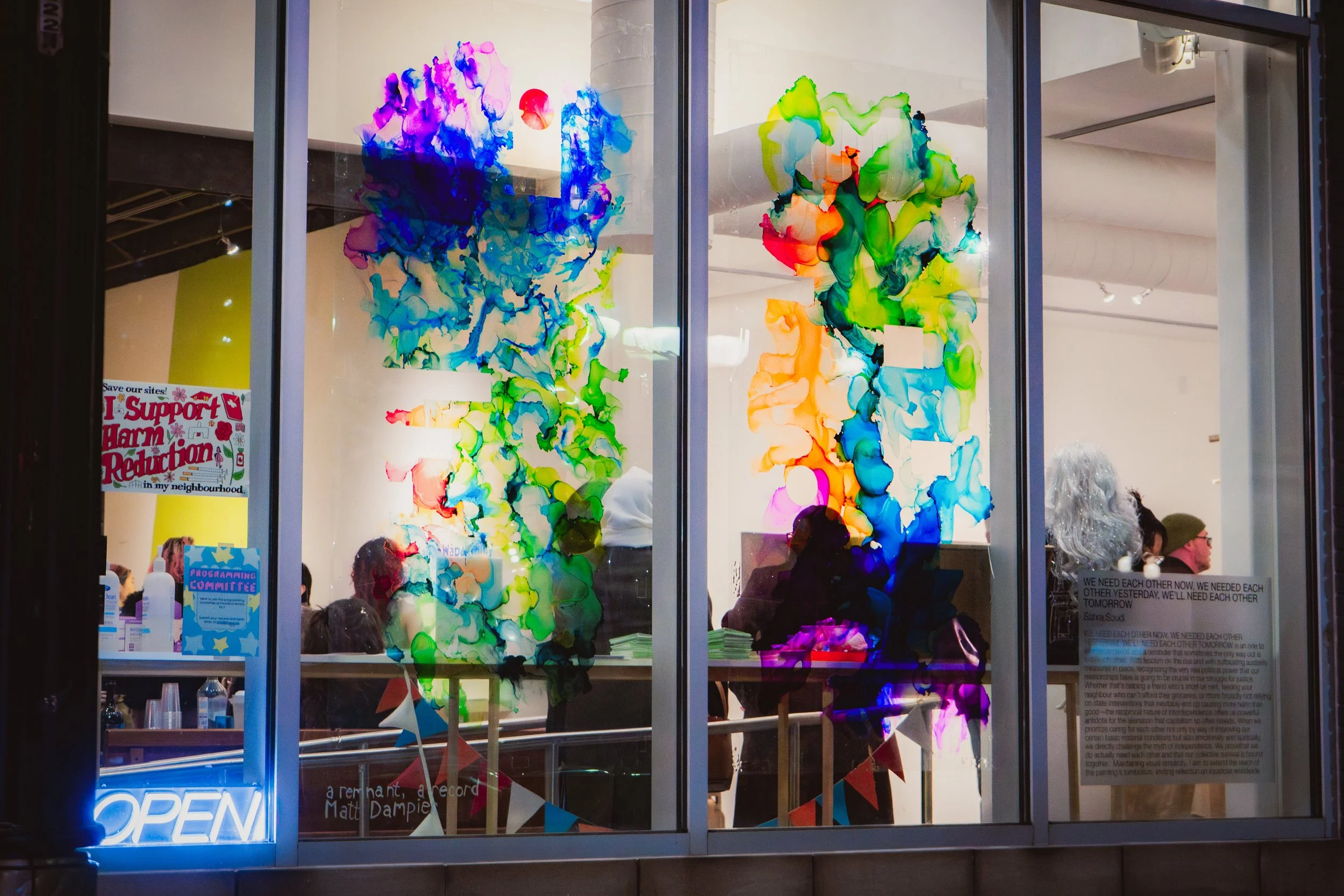 Colourful abstract art on a gallery window with people inside, neon 'OPEN' sign at the bottom left, and various posters and signs on the window.