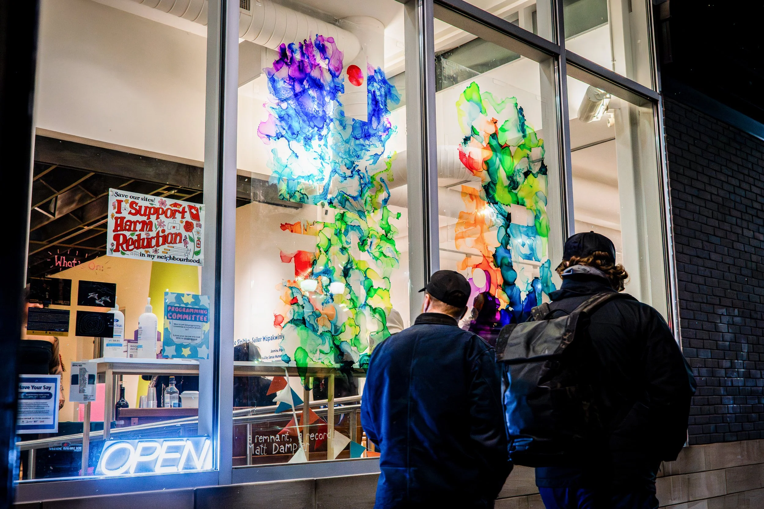 Two people stand outside a colorful storefront window with vibrant, abstract ink art displays. The window features an illuminated 'OPEN' sign, and inside there are signs and supplies.