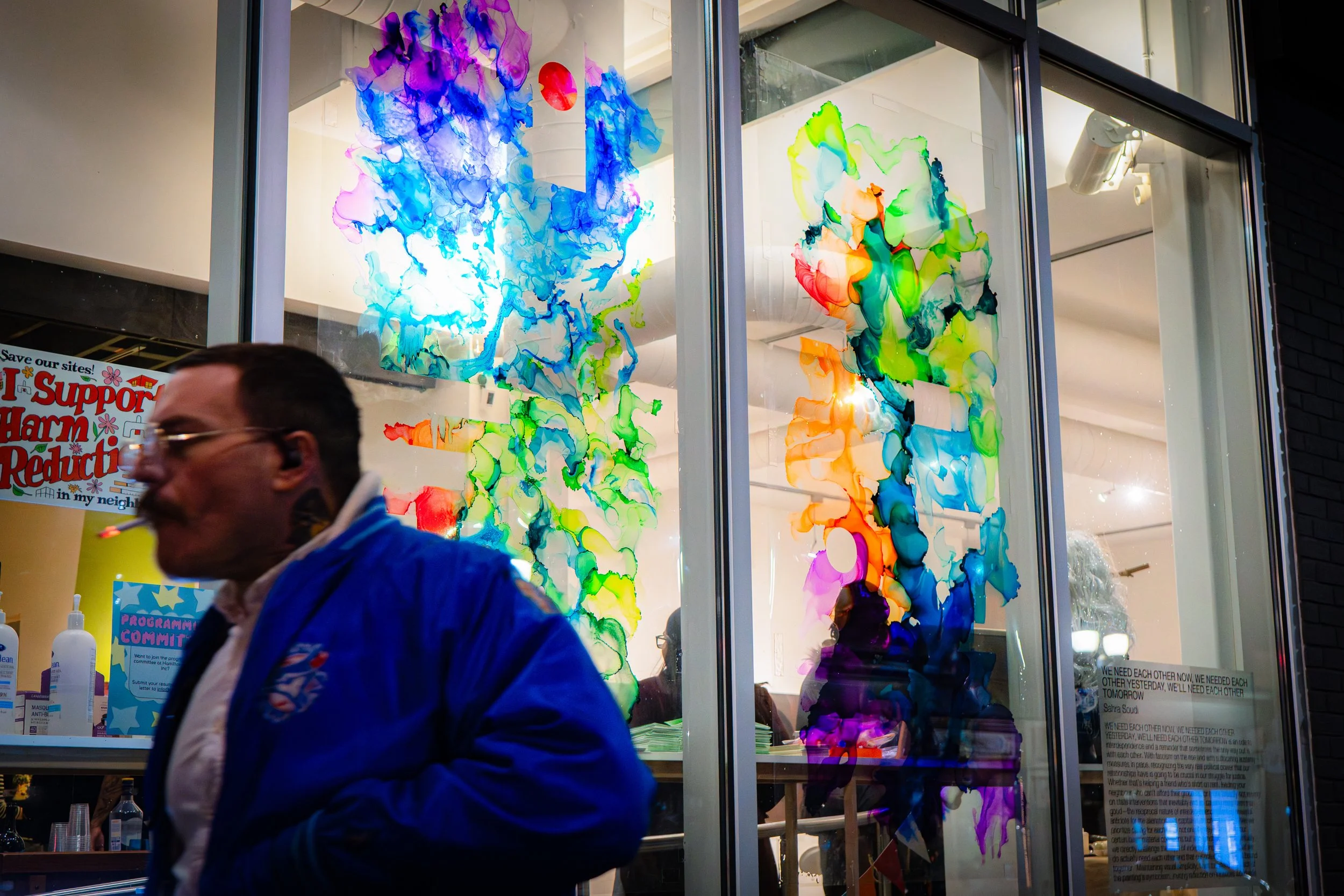 Colorful abstract ink artwork displayed on large glass windows of a building at night, with a man in a blue jacket walking past in the foreground.