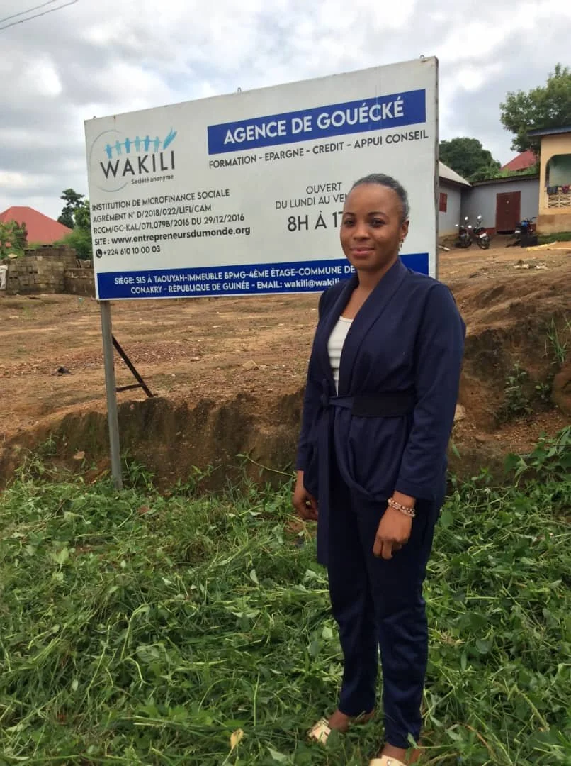 A woman standing outdoors in front of a signboard with informational text. The signboard contains details about an agency named 'Agençe de Gouécké' and is located at a specific address in Guinea.