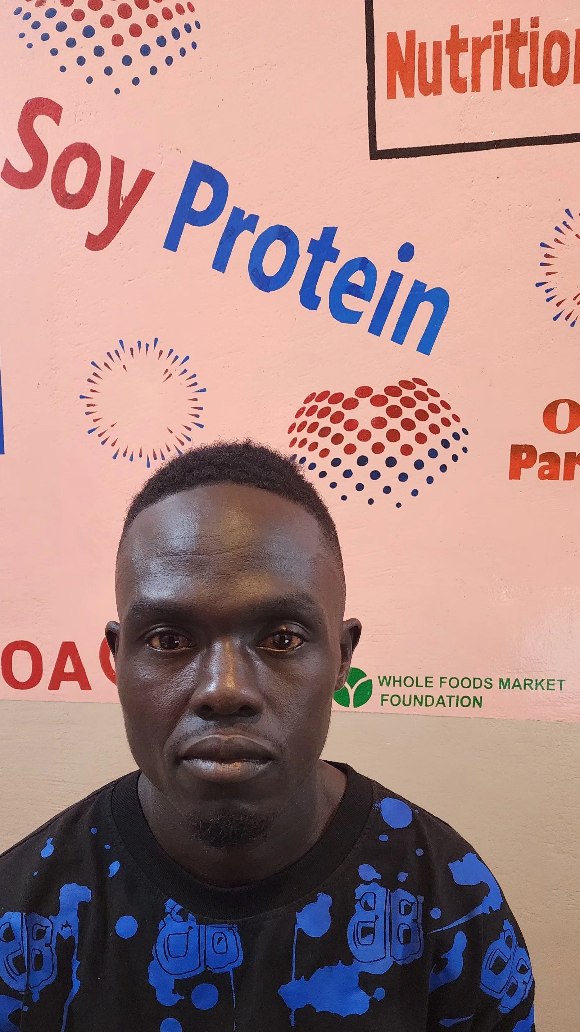 A man with dark skin and short hair looking directly at the camera, wearing a black shirt with blue patterns. Behind him is a pink wall with text about nutrition and whole foods, including the phrases 'Soy Protein' and information about the Whole Foods Market Foundation.