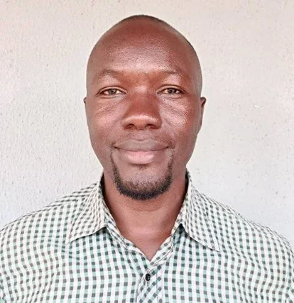 A man with a shaved head and goatee wearing a checkered shirt standing against a plain background.