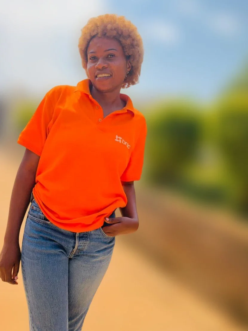 A woman with short curly blonde hair wearing an orange polo shirt and light blue jeans standing outdoors with a blurred background of sky and greenery.