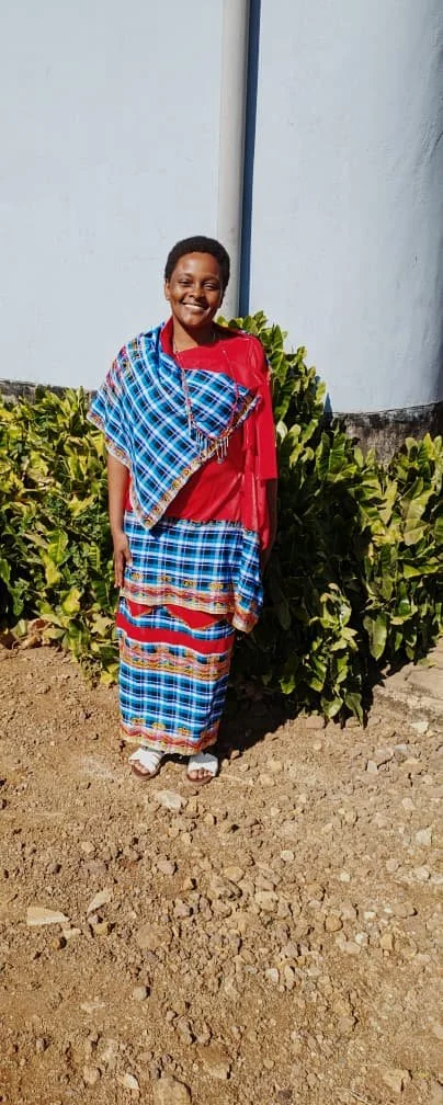 A young girl wearing traditional Maasai clothing, including a red top and blue patterned shuka, standing outdoors in front of green shrubs and a white building wall.