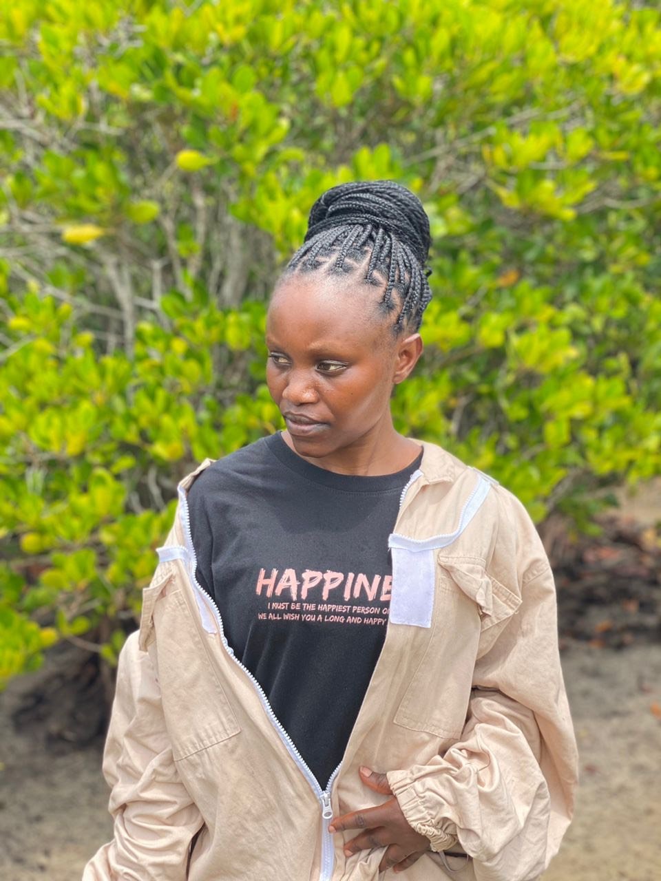 A woman with braided hair wearing a black t-shirt with the word 'HAPPINESS' and a beige jacket, standing outdoors in front of green bushes.