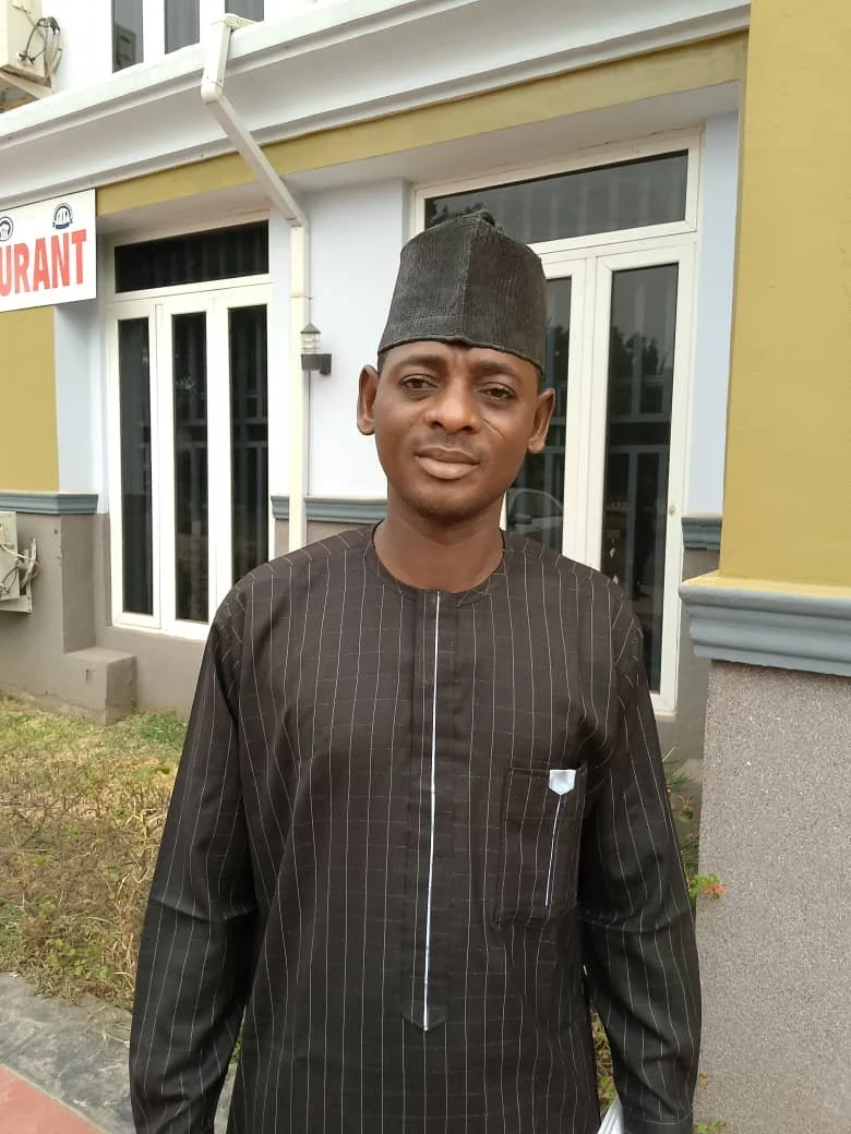 A man wearing a dark traditional Nigerian outfit and a black cap, standing outside near a building with white-framed windows and a yellow wall.