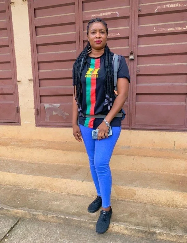 A woman standing outdoors in front of a reddish-brown wooden gate, wearing a black T-shirt with colorful stripes and a lightning logo, blue jeans, and black sneakers, holding a smartphone in her right hand.