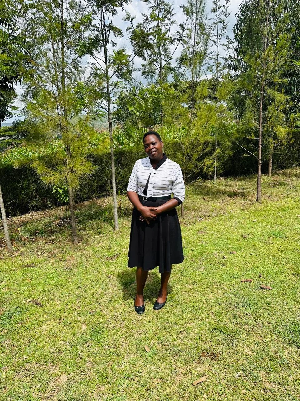A woman in a black skirt and striped blouse standing on grass with green trees in the background.