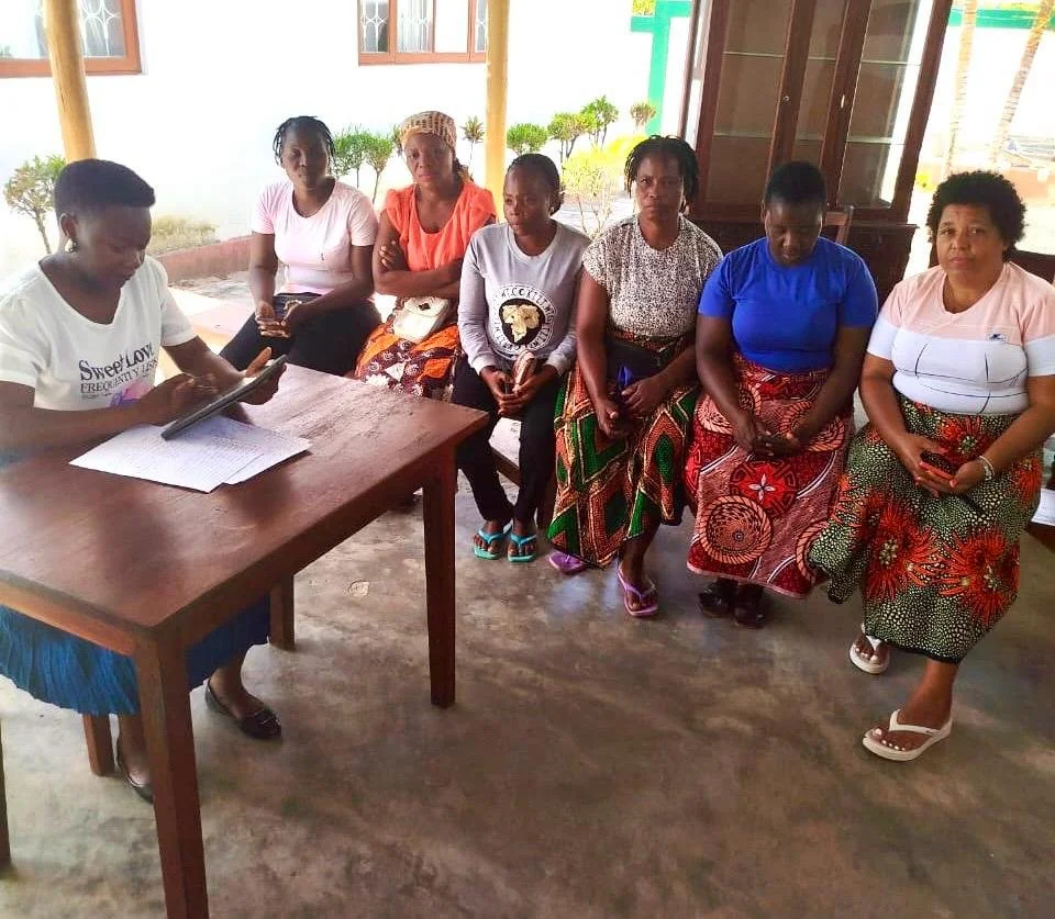 Seven women sitting indoors near a wooden wall, with one woman sitting at a table looking at a tablet, while the other six women sit in a row, some looking at their phones, dressed in colorful skirts and casual tops.