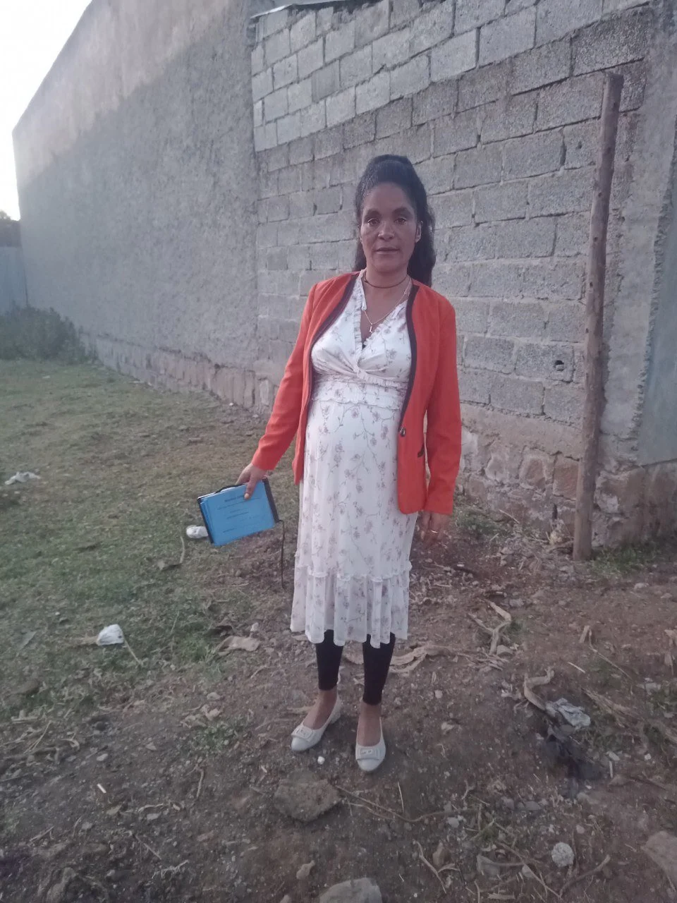 A woman standing outdoors on dirt ground with a brick wall behind her, holding a small blue book or notebook.