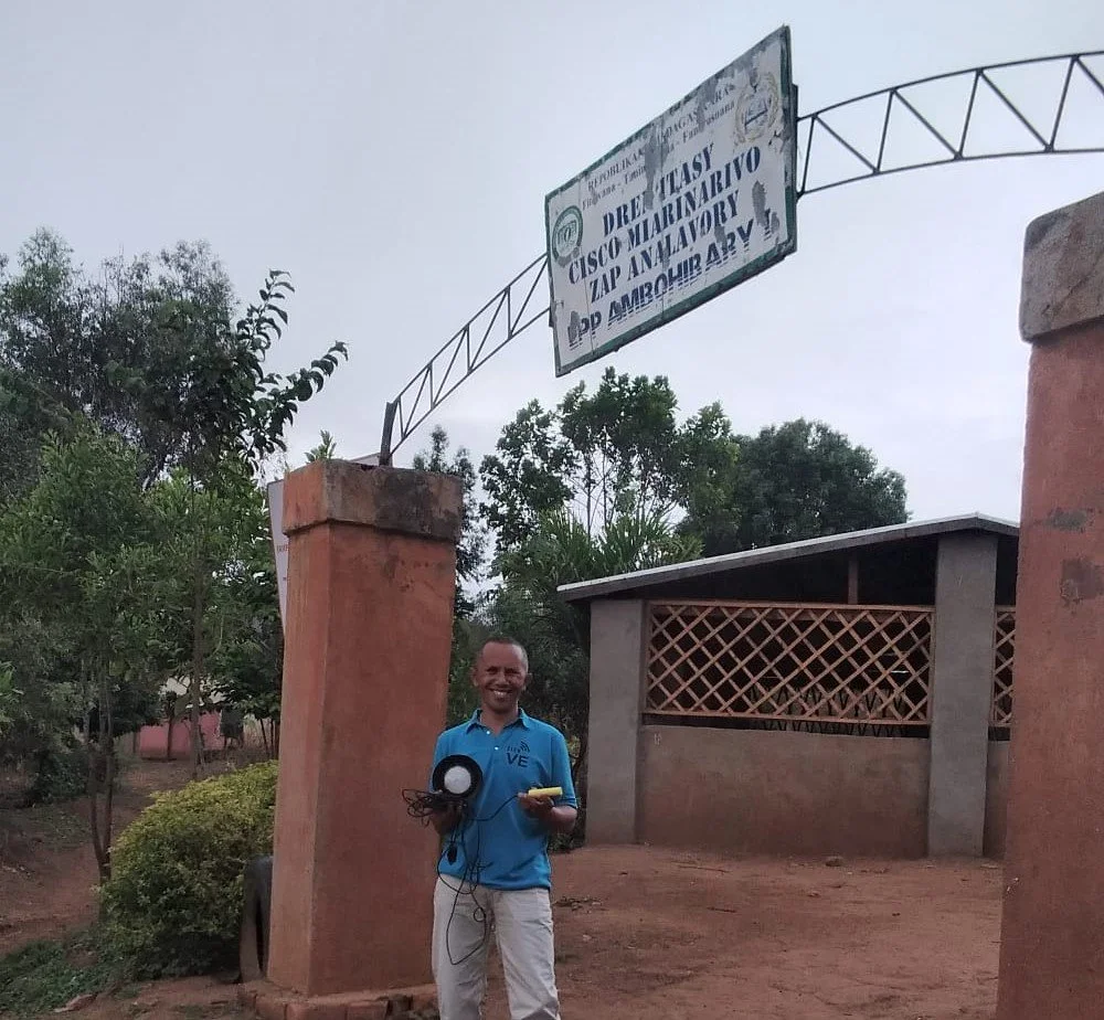 Man standing outdoors near a brick pillar and a building, holding a microphone and a yellow device, smiling, with a signboard hanging above.