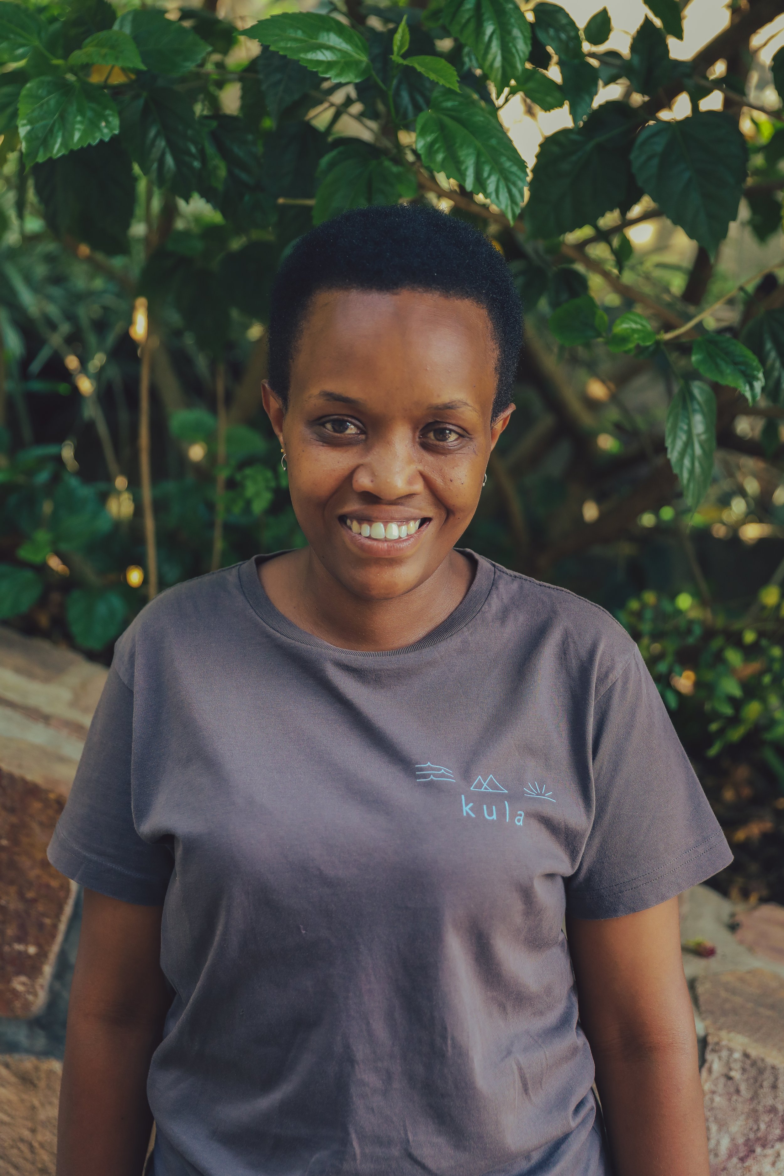 A woman with short black hair smiling outdoors, wearing a gray T-shirt with a minimalistic mountain, wave, and sun design and the word 'kula' on it, standing in front of green foliage.