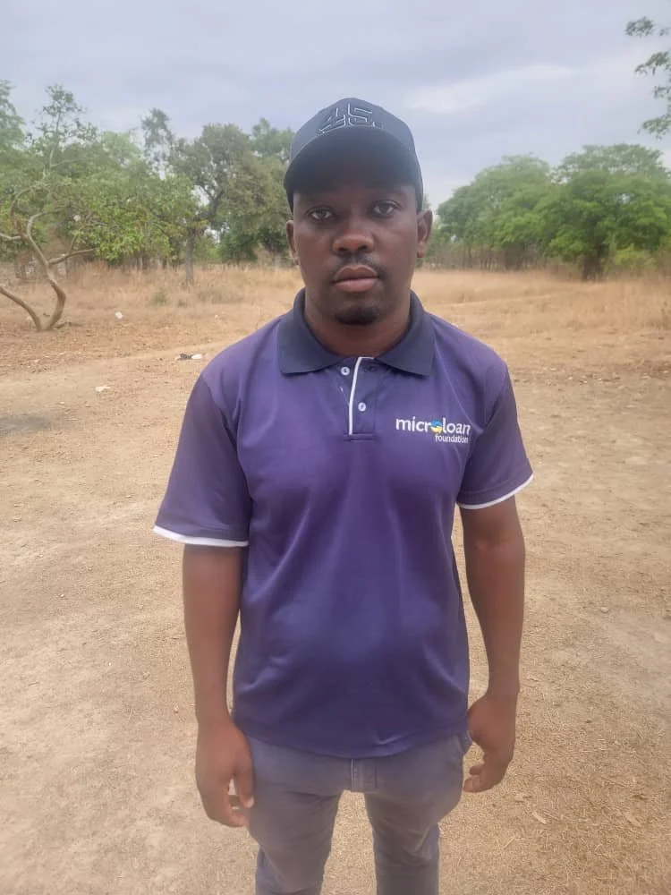 A young man standing outdoors in a dry, open area with scattered trees, wearing a purple polo shirt with a logo and a black cap, looking directly at the camera.