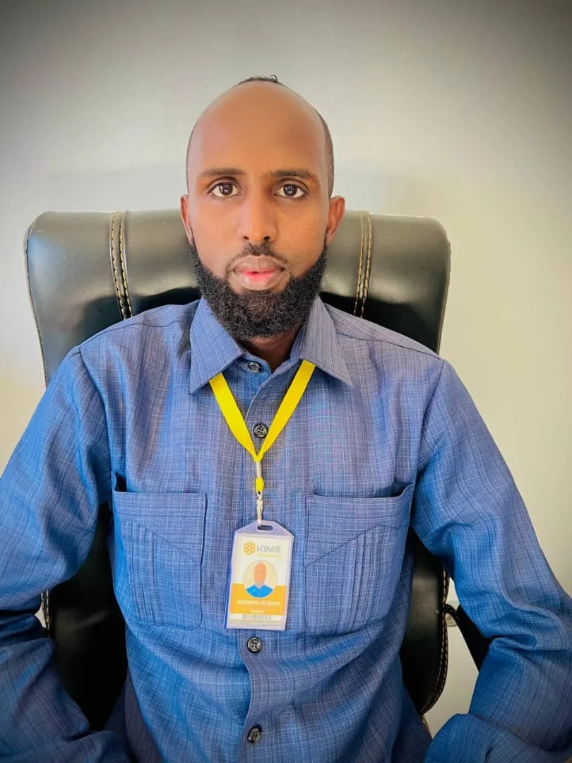 A man with a beard sitting in a black office chair, wearing a blue checkered shirt and a yellow lanyard with an ID badge. The badge reads 'KIMS' and has a photo and title.
