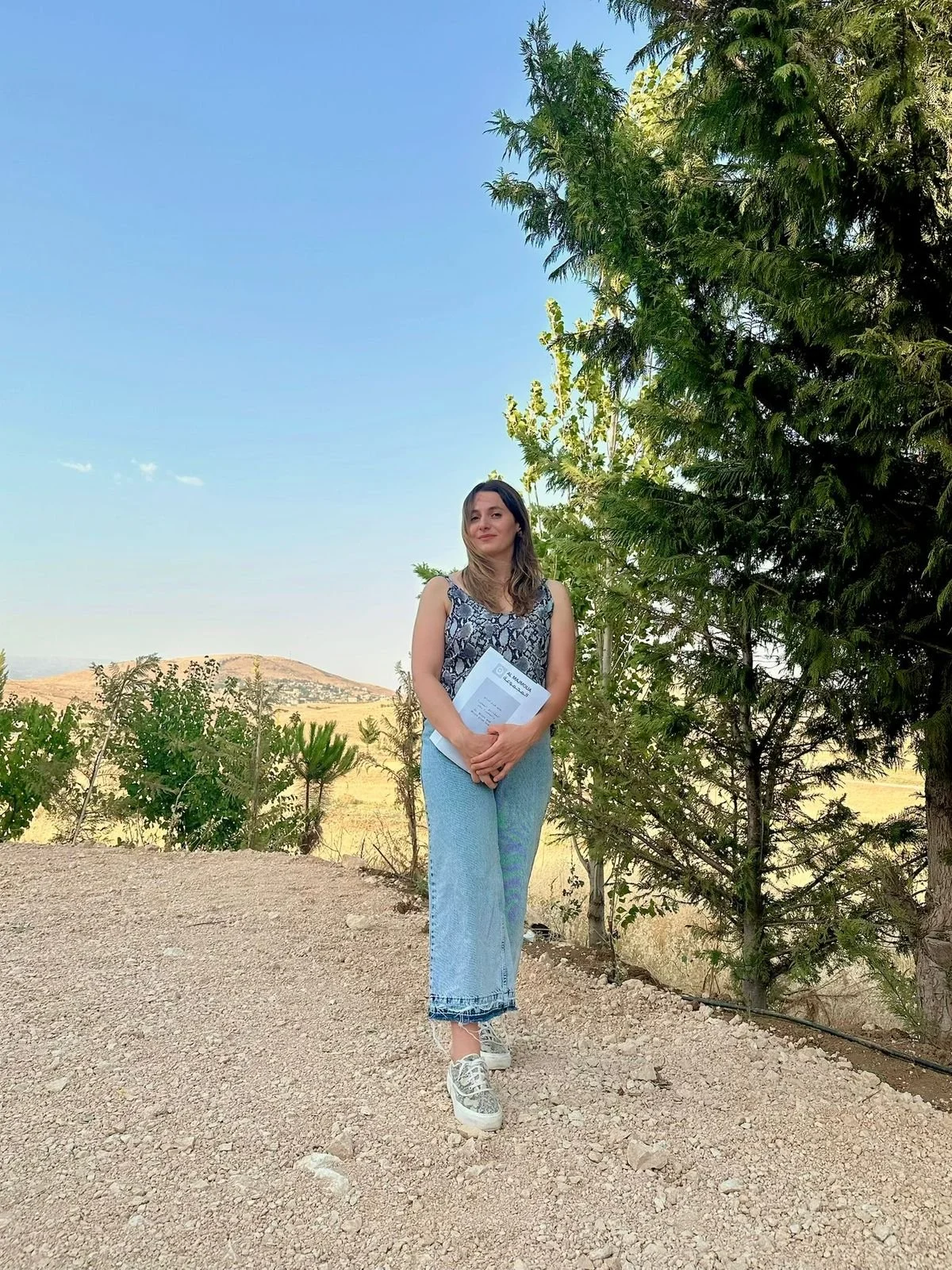 A woman standing outdoors on a dirt path holding a folder, with trees and hills in the background under a clear blue sky.