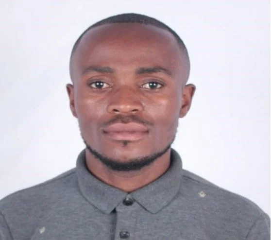 A young man with short hair and a goatee, wearing a gray collared shirt, standing against a plain white background.