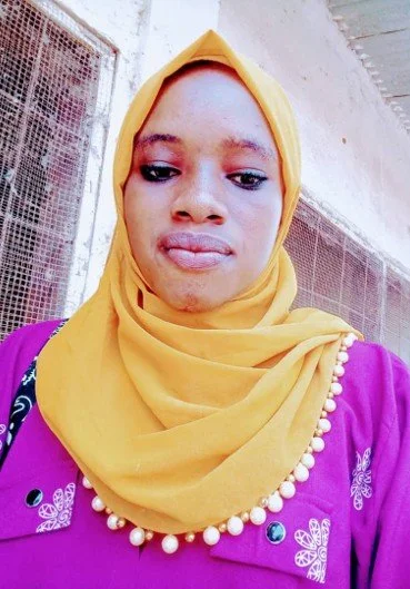 A young woman wearing a yellow headscarf and a purple top with embroidered butterfly patterns, standing outdoors near a building with a window.